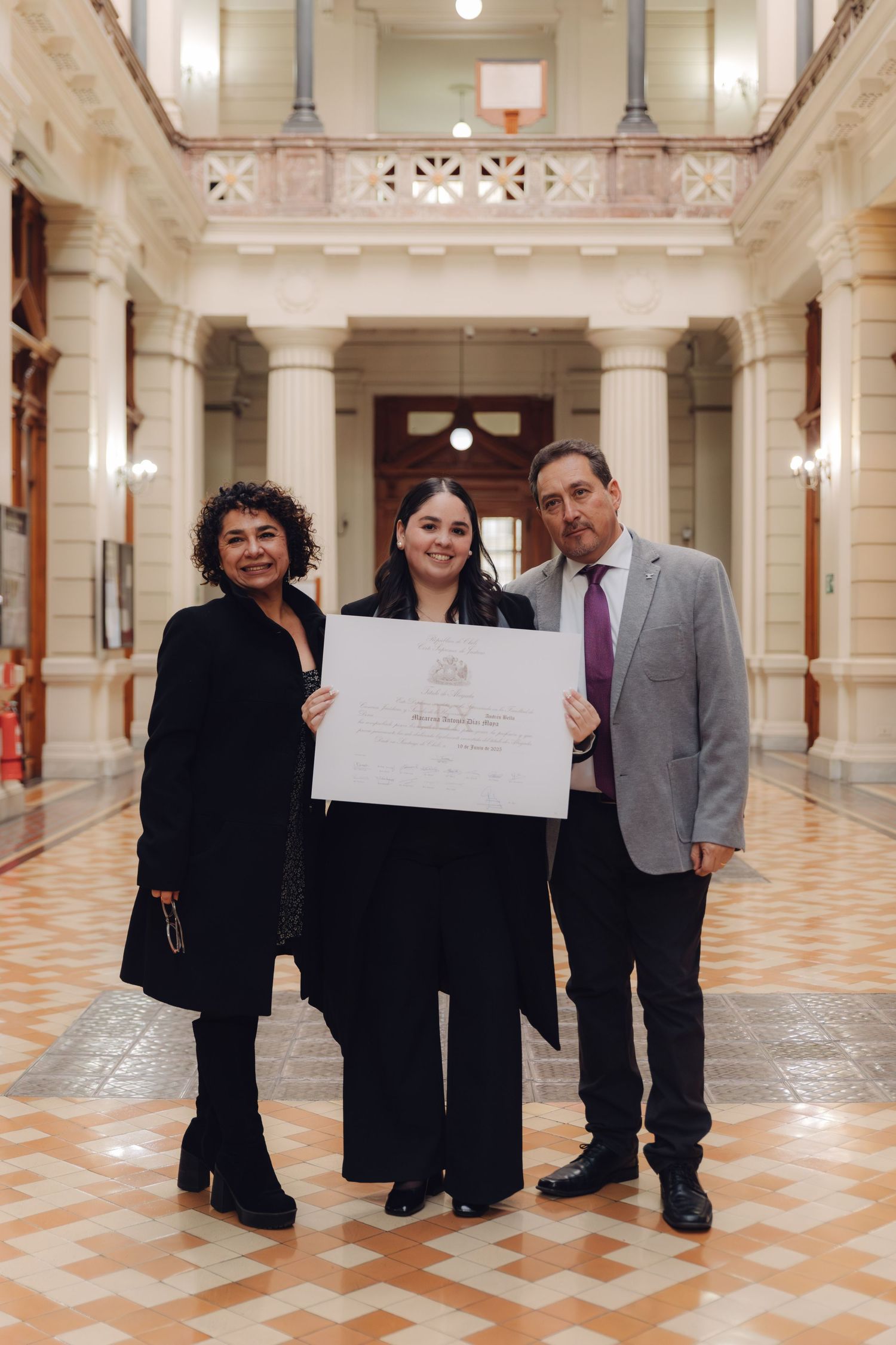 Graduate celebrates with parents holding diploma in classic architectural hall.