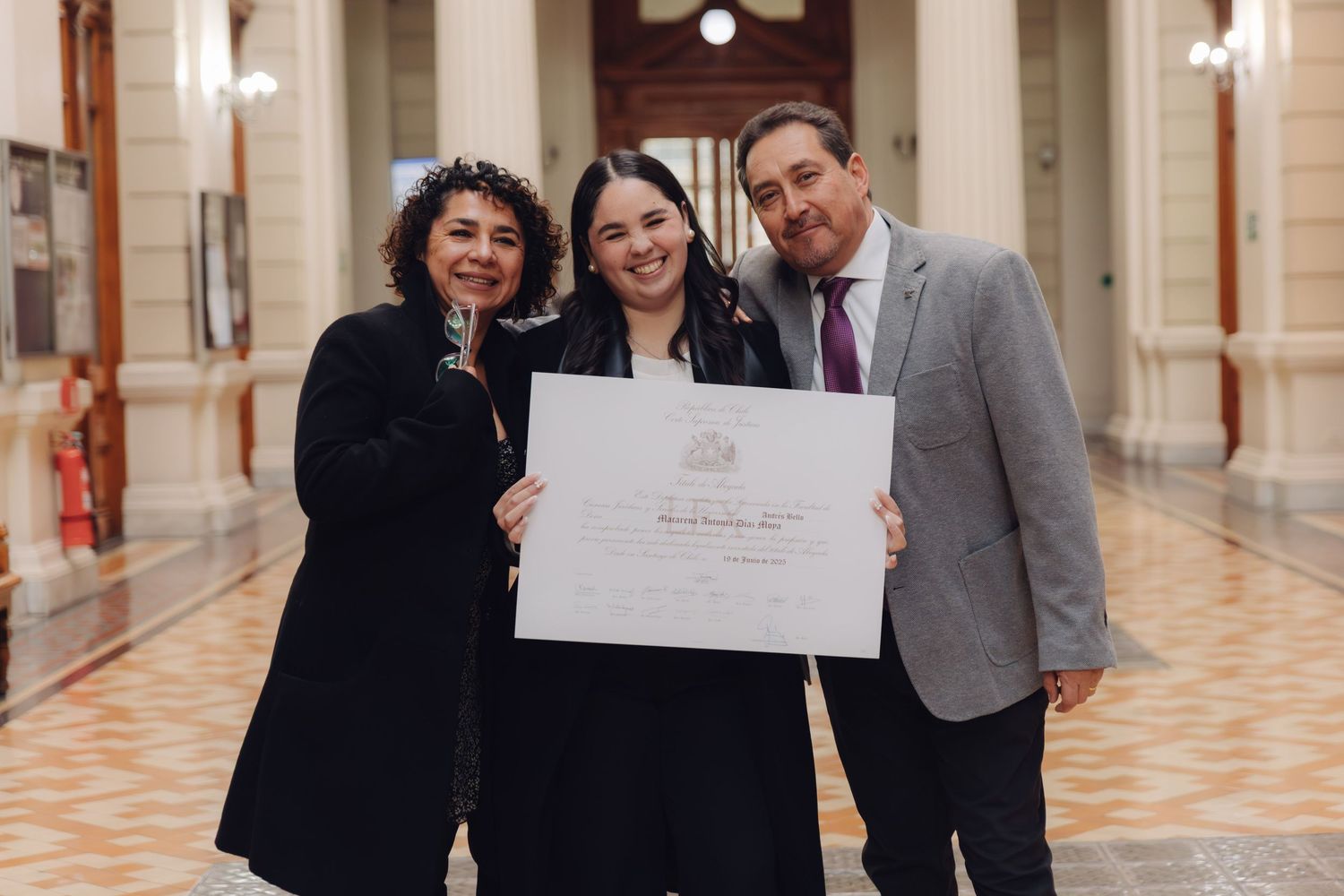 Proud graduate poses with family displaying official diploma in formal venue.