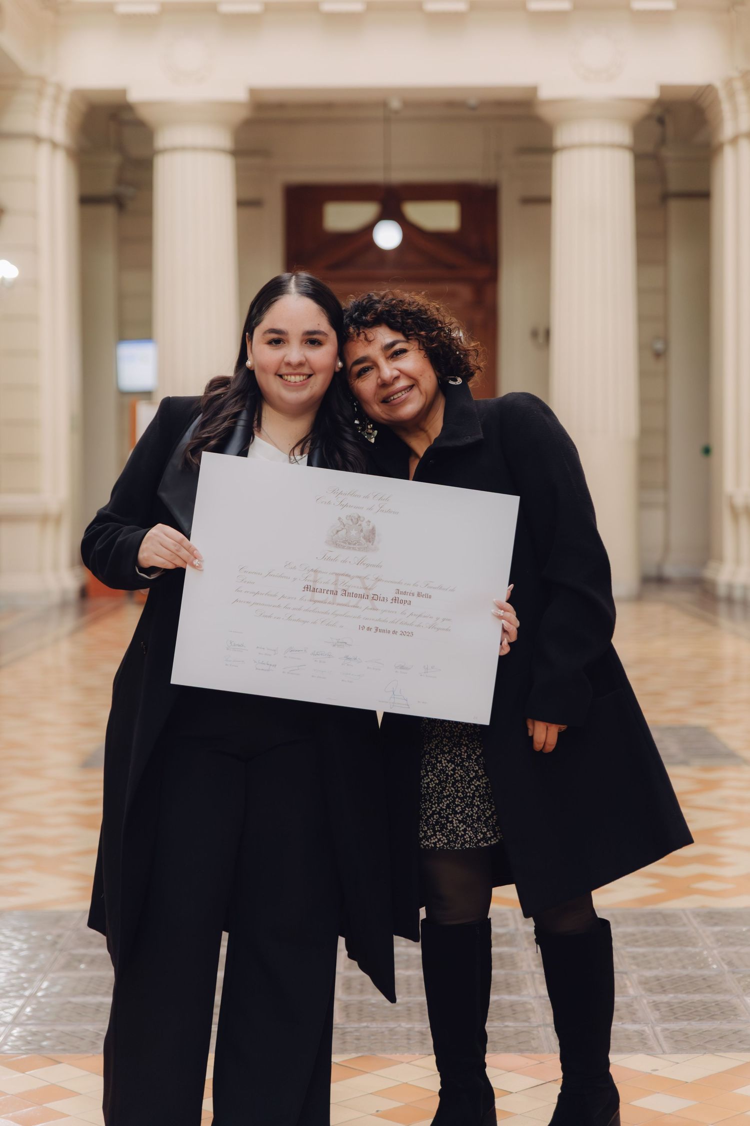 Two women smile together holding graduation certificate in columned building.