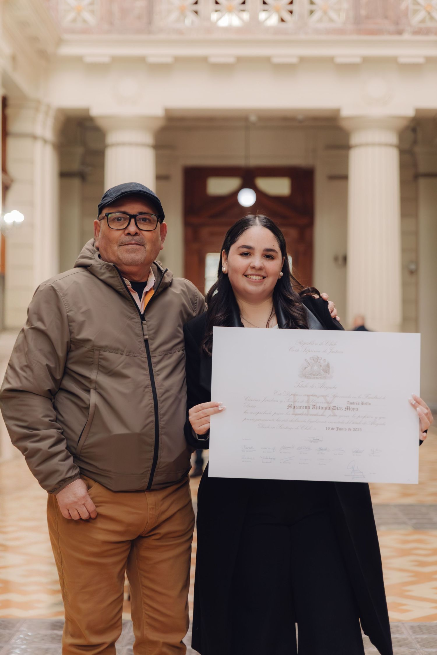 Young graduate and older man proudly display official diploma in hall.