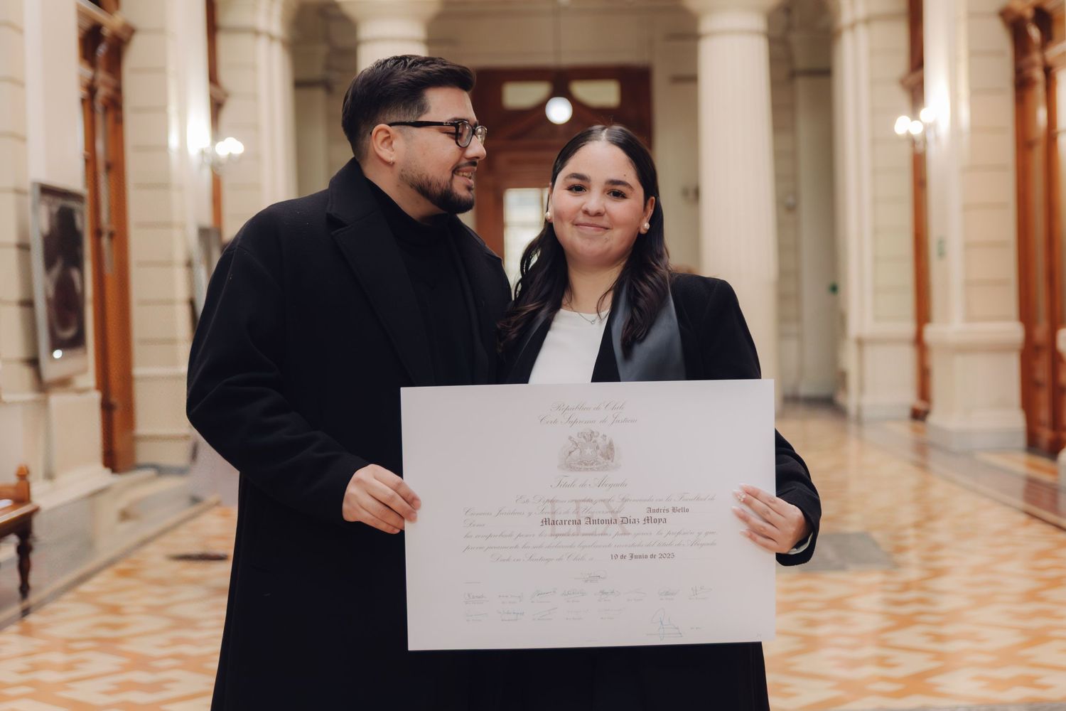 Man and woman sharing a joyful moment while holding a graduation diploma in a classic hallway.