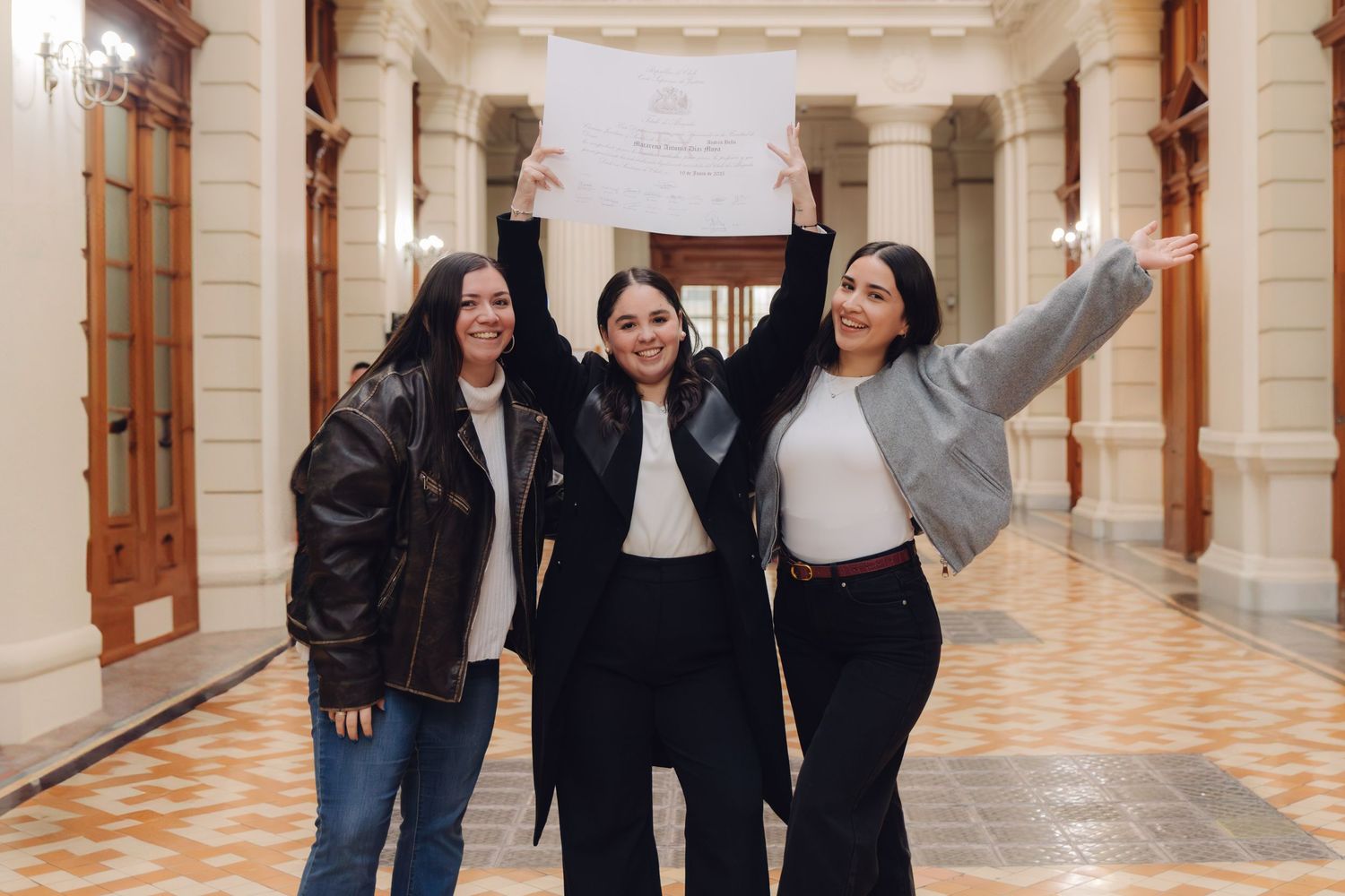 Three women celebrating joyfully while holding up a certificate in a bright elegant corridor.