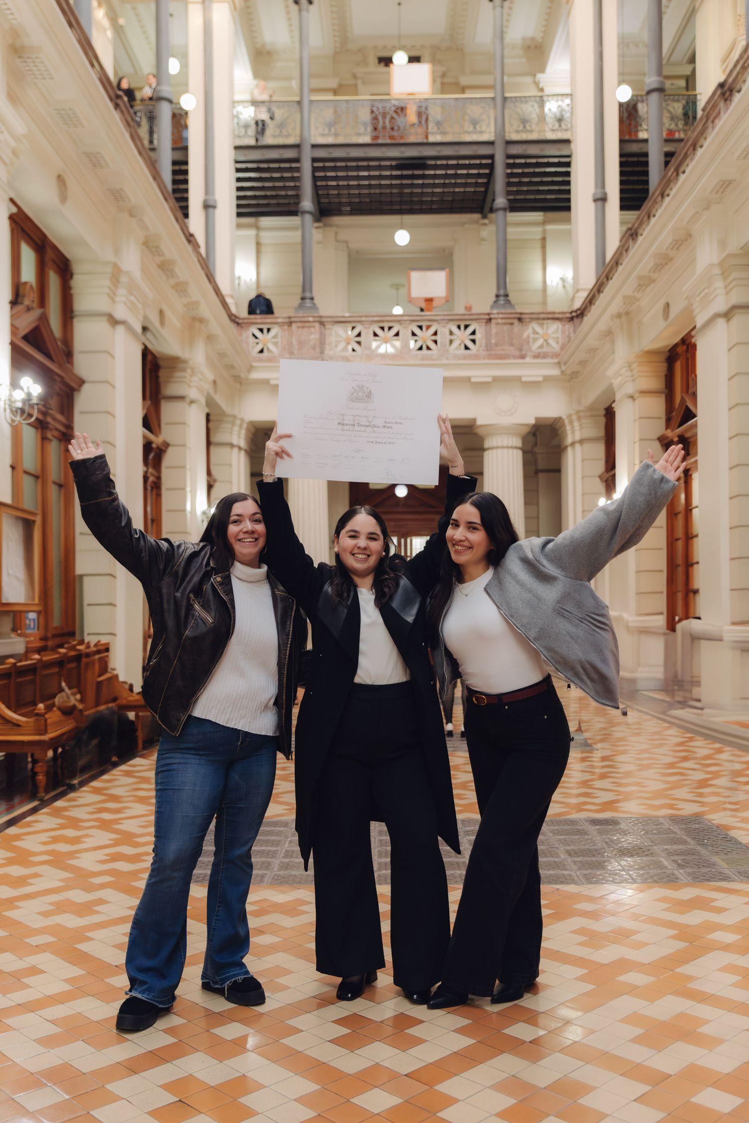 Three women cheering and raising a graduation certificate high in a grand historic building interior.