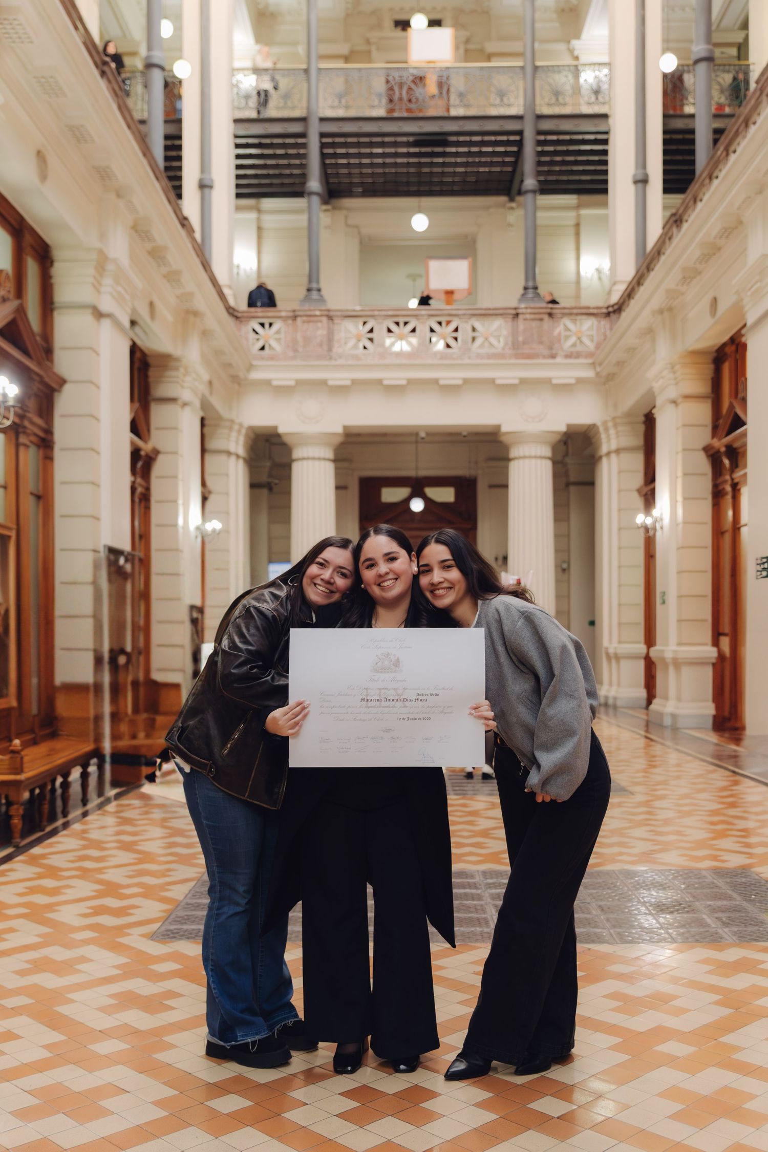 Three smiling women posing together holding a graduation diploma in a beautiful historic hallway.