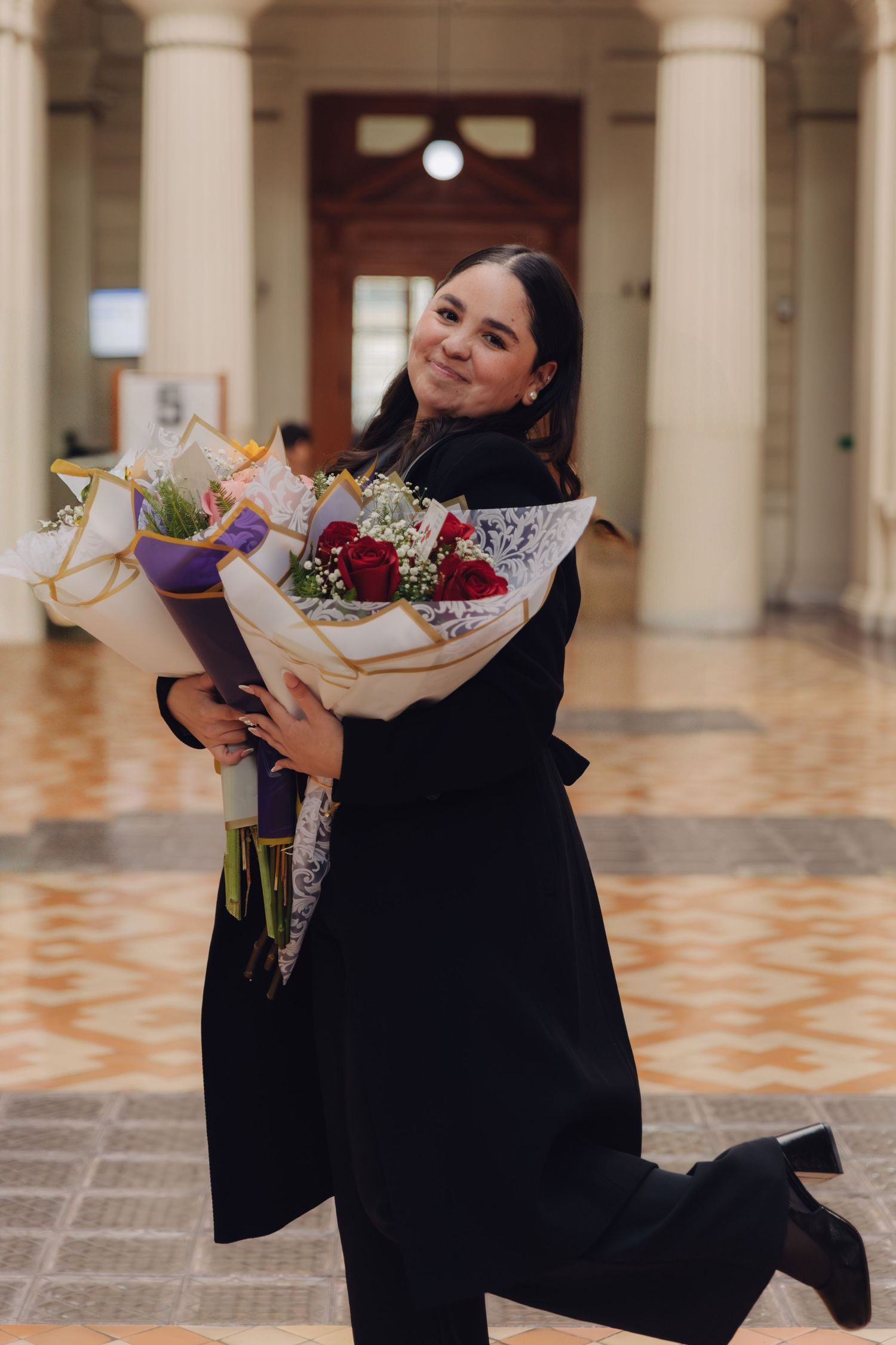 Smiling graduate in black coat holding a bouquet of red roses in an elegant tiled hallway.