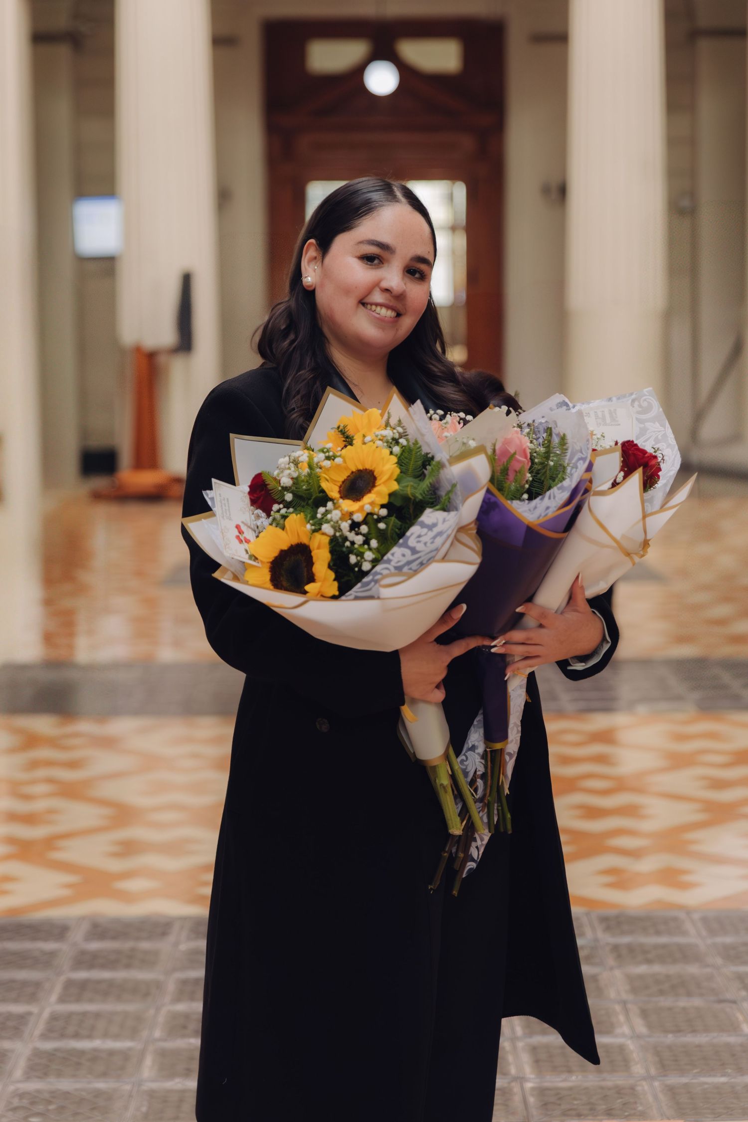 Happy graduate holding colorful flower bouquets including sunflowers in a grand classical building.