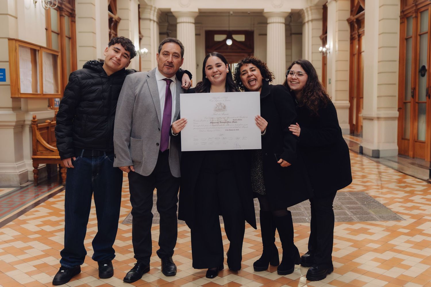 Family of five celebrating together while holding a graduation diploma in a classic hallway.