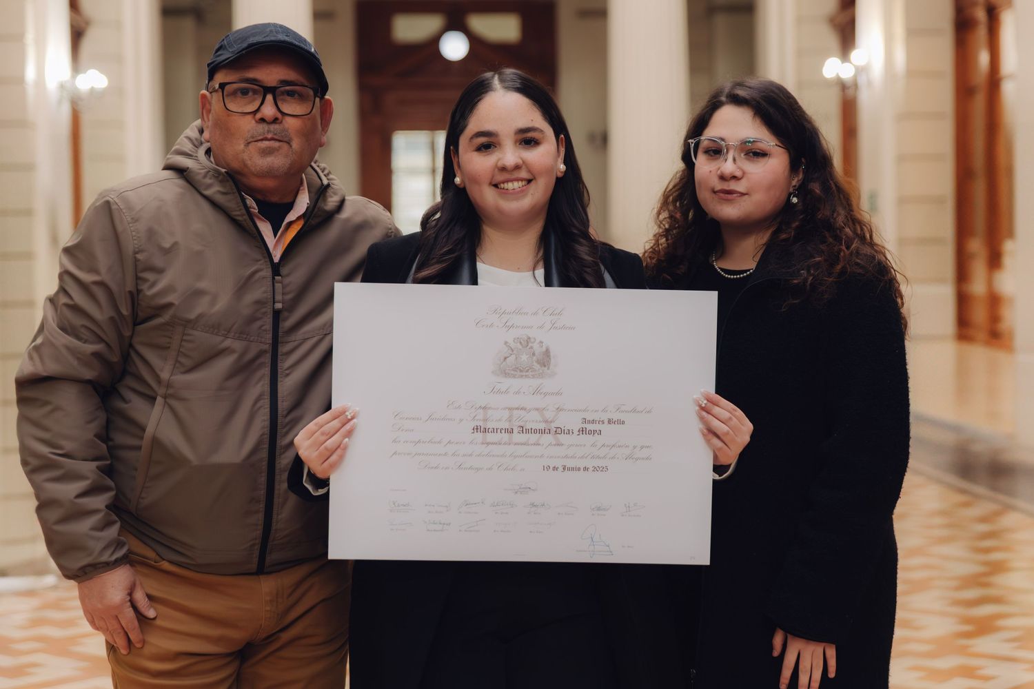 Graduate proudly holding a diploma flanked by two family members in an elegant corridor.