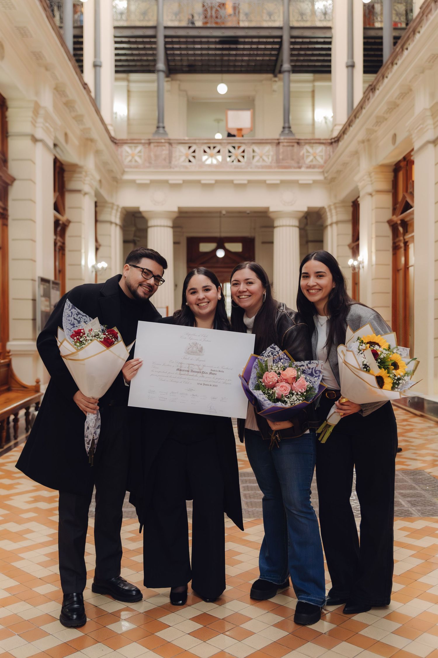 Four people celebrating a graduation indoors, holding flowers and a diploma certificate in an elegant building.