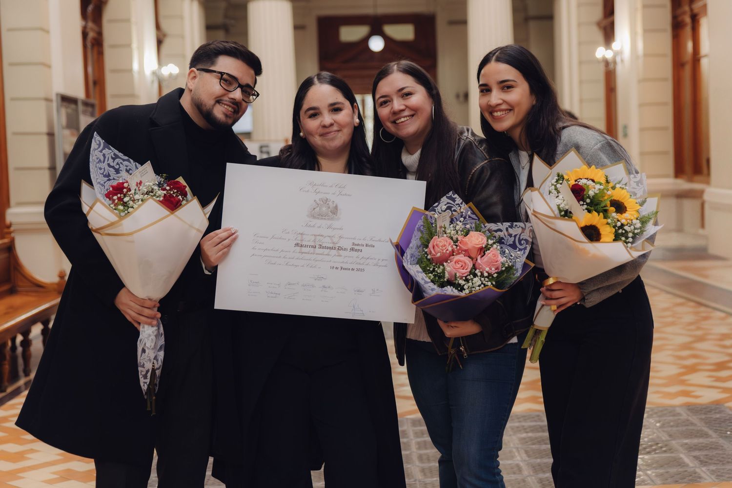 Group of four smiling graduates posing with flowers and a diploma certificate at a graduation ceremony.