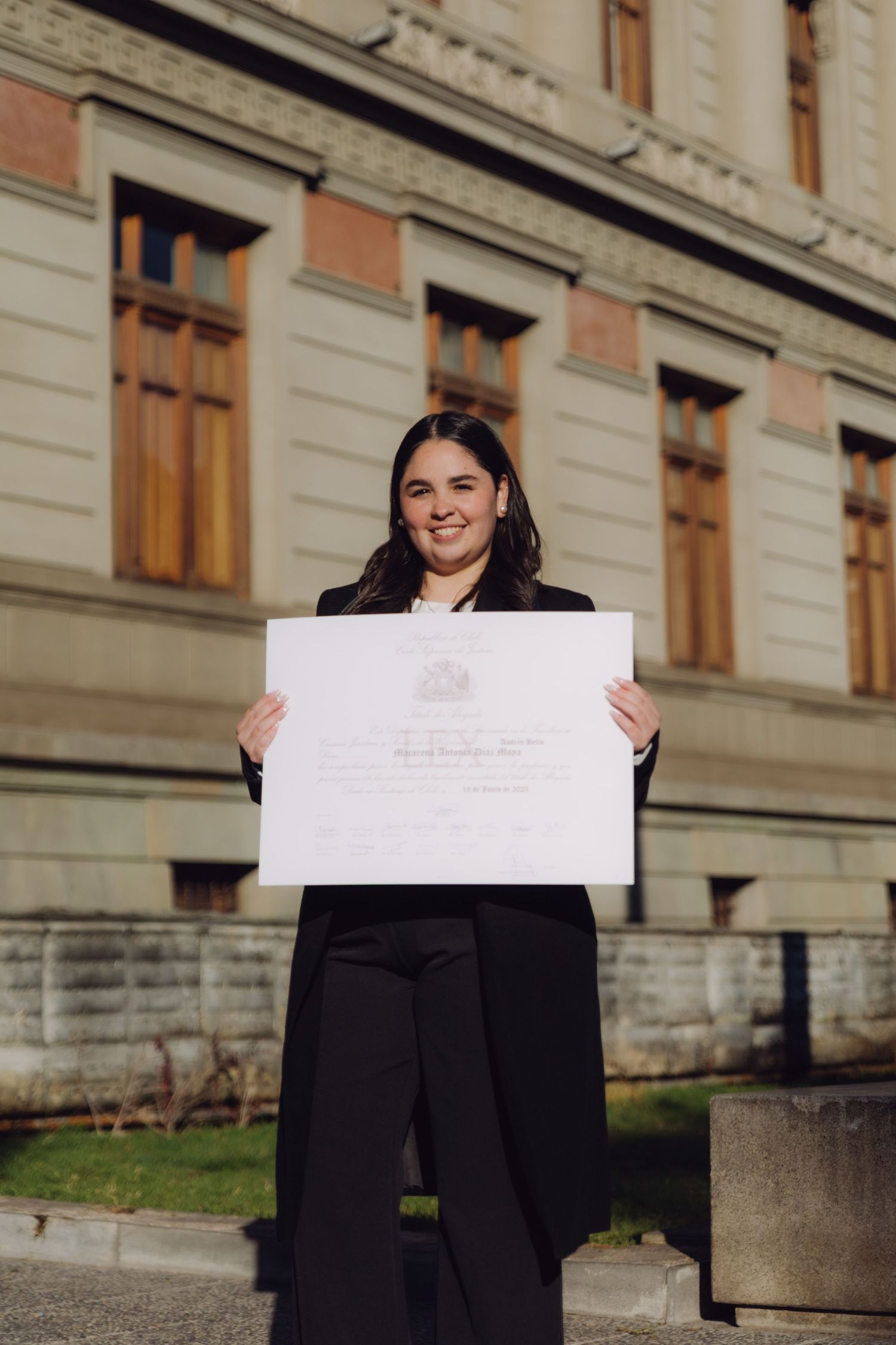 Young woman in black blazer proudly holding her diploma certificate outside a historic university building.