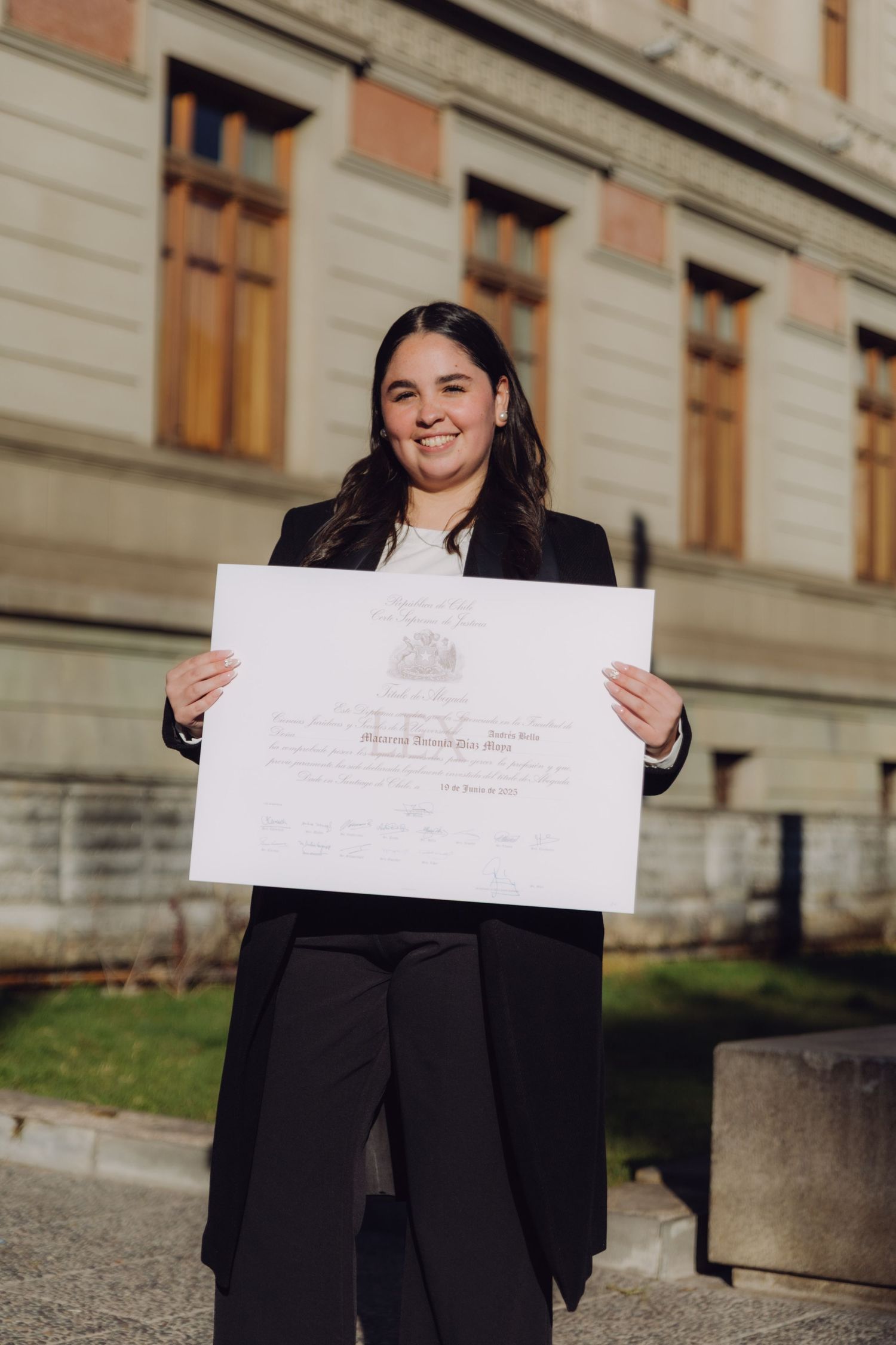 Smiling graduate in black blazer displaying her diploma certificate outdoors at a university campus.