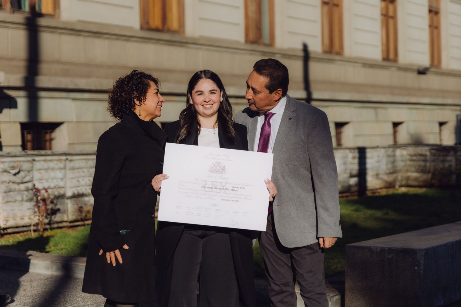 Graduate sharing diploma celebration moment with two proud family members outside a historic building.