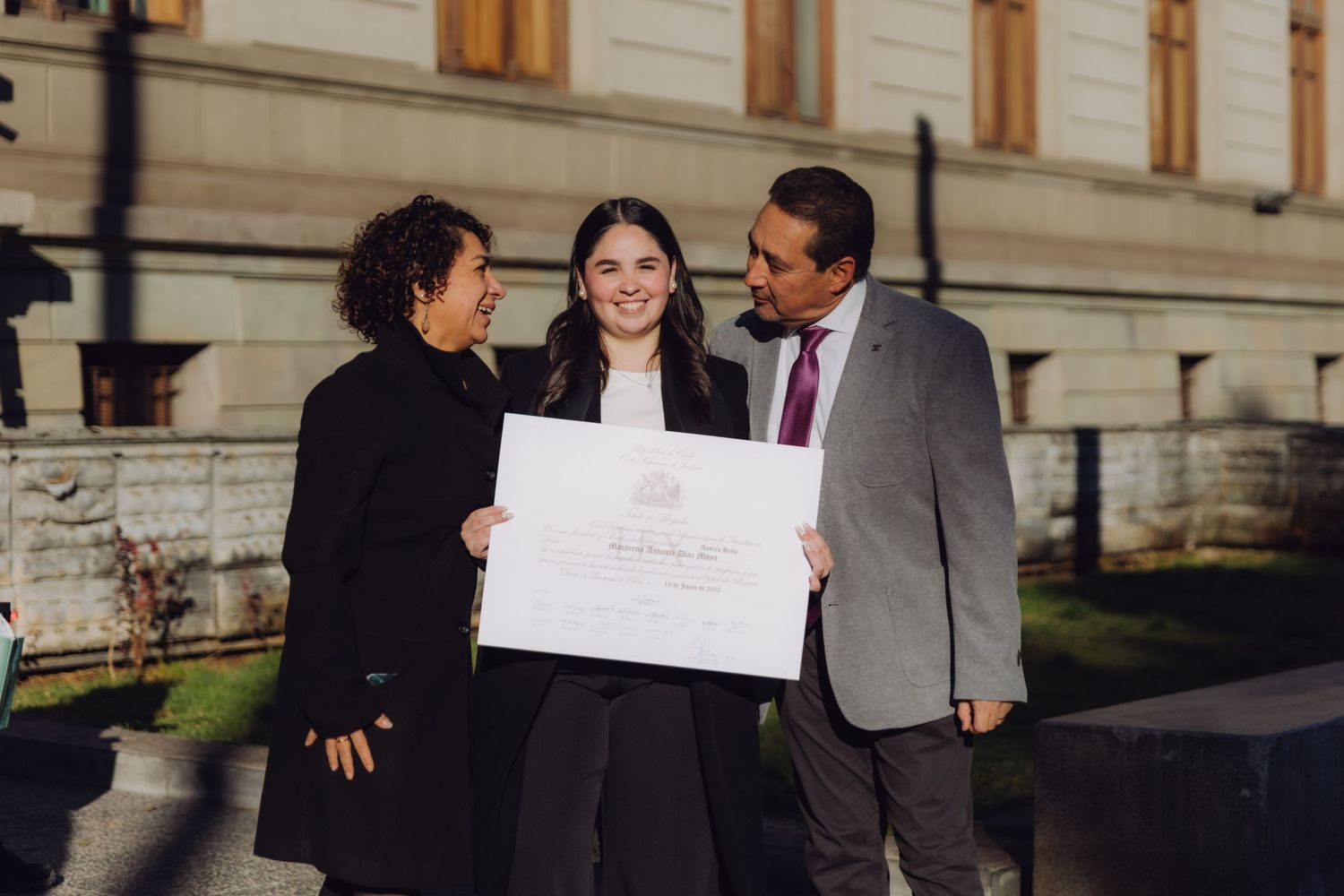 Young graduate posing with her parents while holding a diploma certificate outside a university.