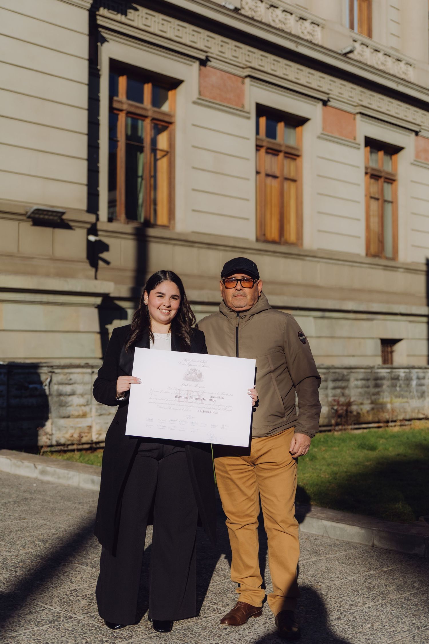 Graduate and older man proudly displaying a diploma certificate together outside a stone building.