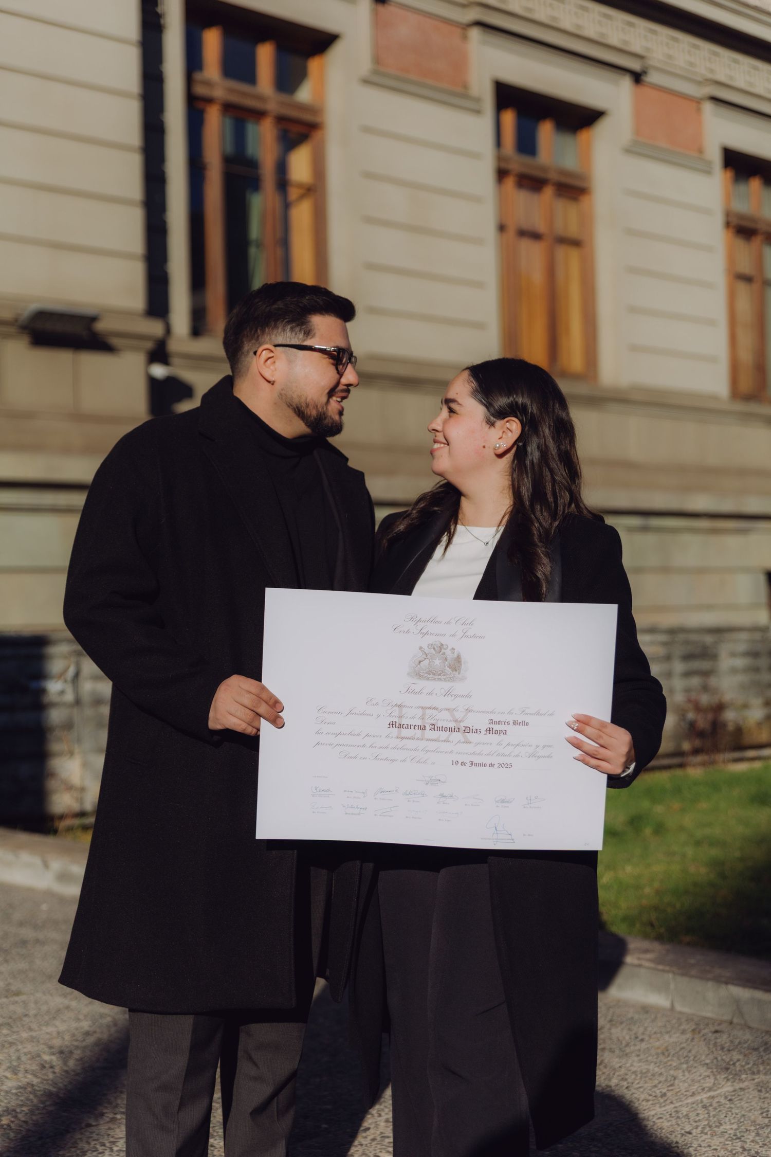 Two people in black coats sharing a joyful moment while holding a graduation diploma certificate.