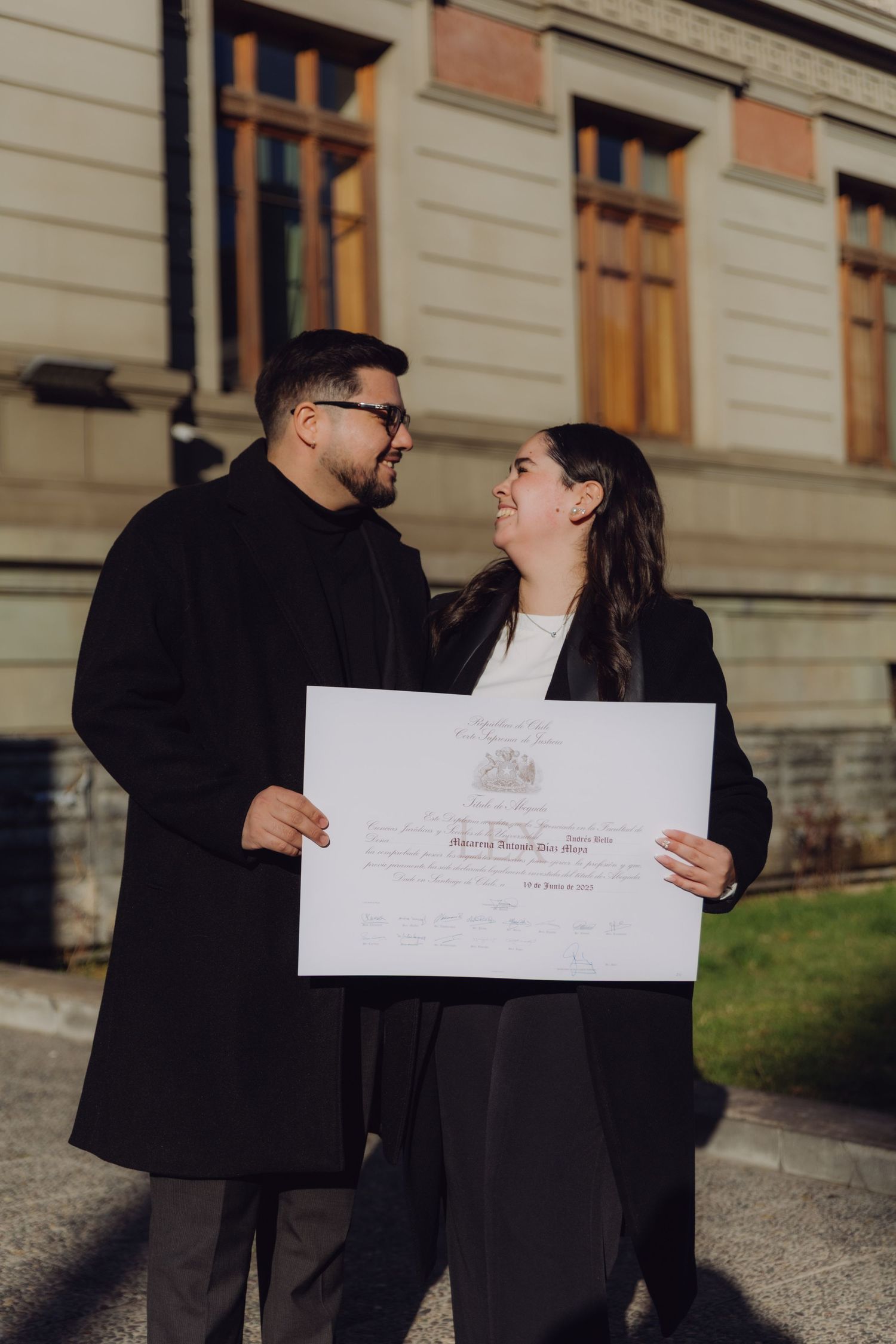 Couple in matching black coats smiling at each other while holding a graduation diploma outside.