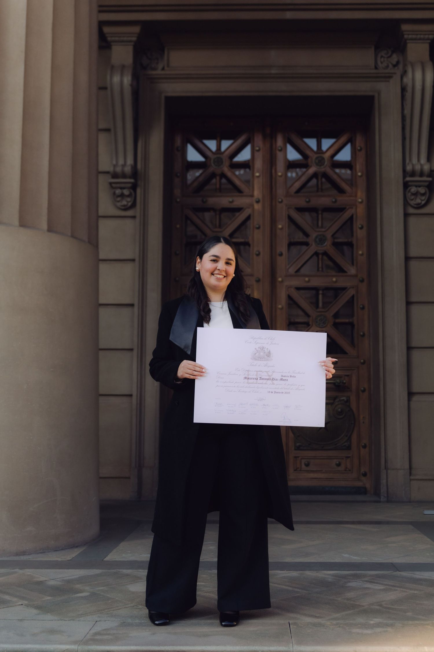 Young woman in black graduation gown proudly holding diploma certificate outside ornate building doors.