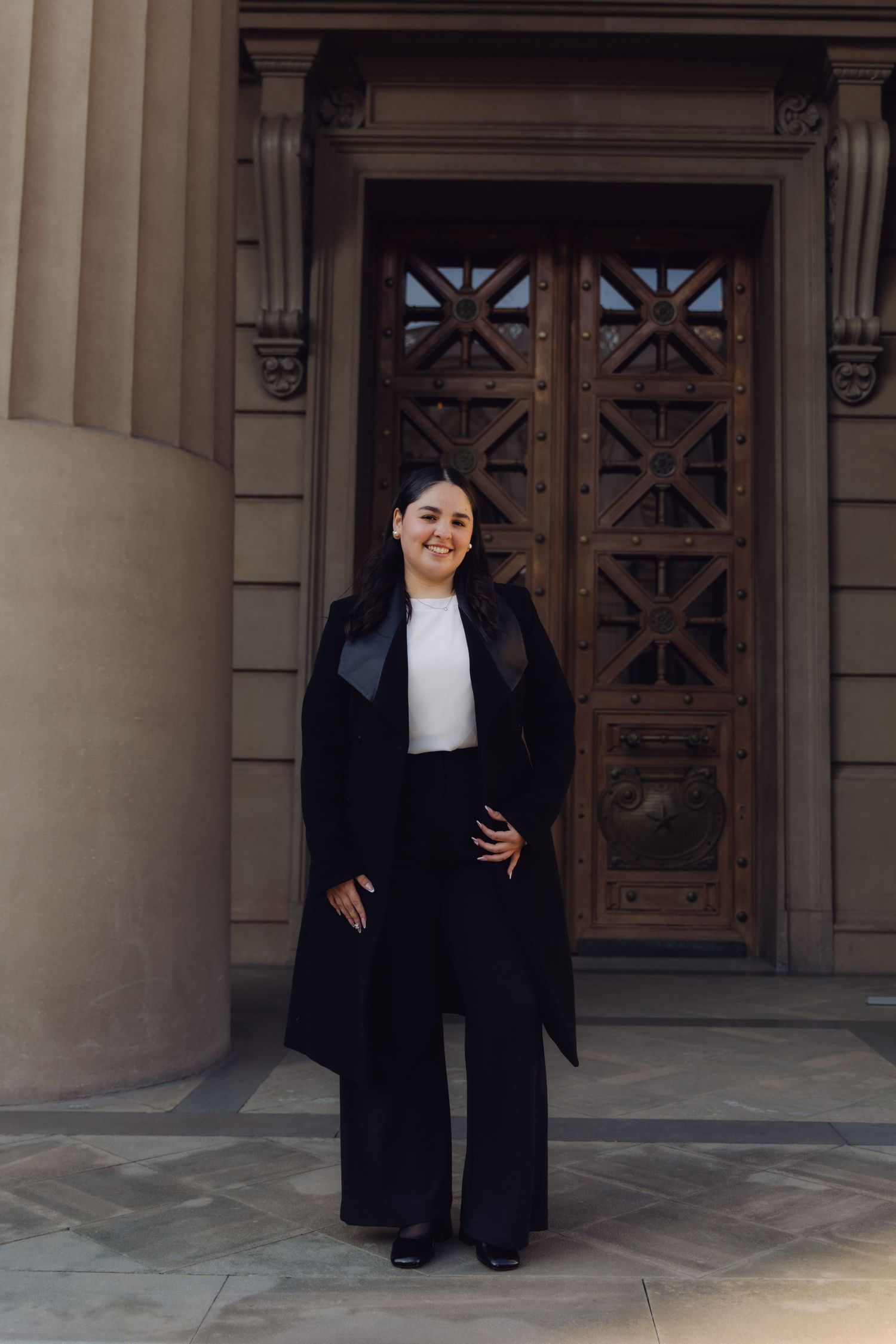 Smiling young woman in black coat and wide-leg pants posing before decorative wooden doors.