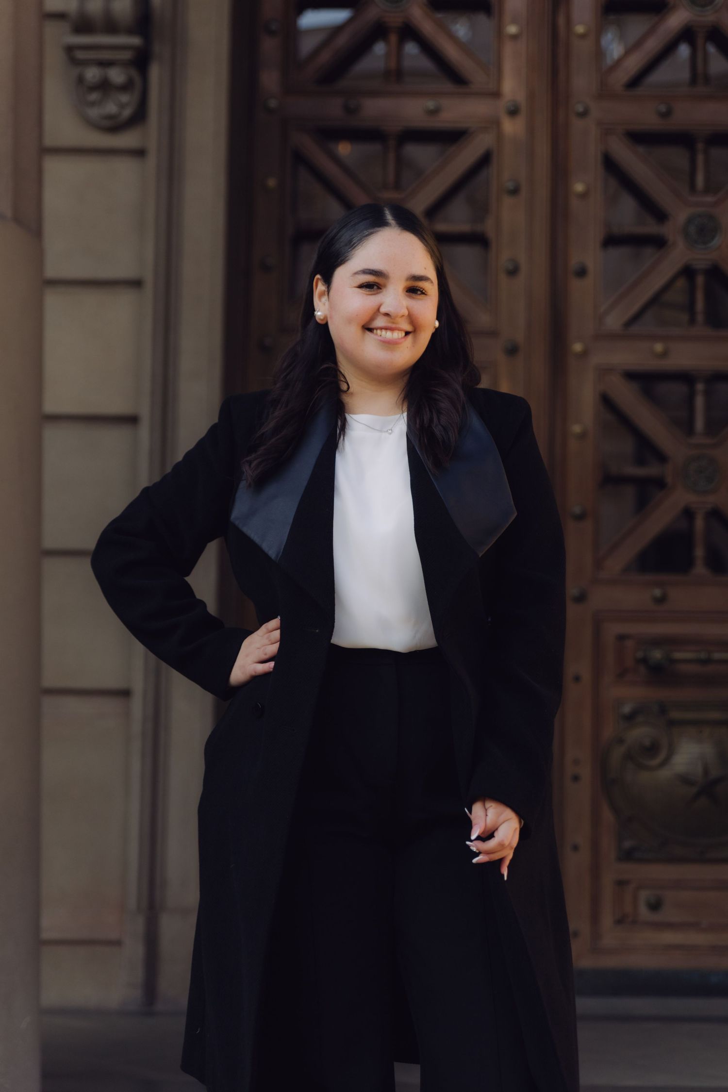 Confident young woman in black blazer coat with white top smiling outside historic building entrance.