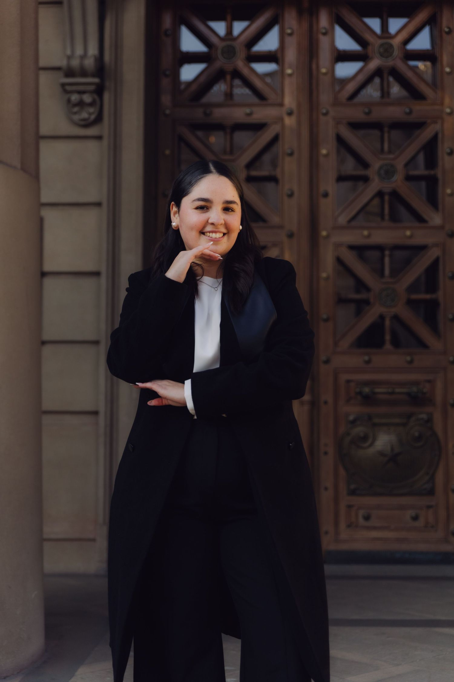 Young woman in black graduation attire posing thoughtfully with hand on chin near ornate wooden doors.