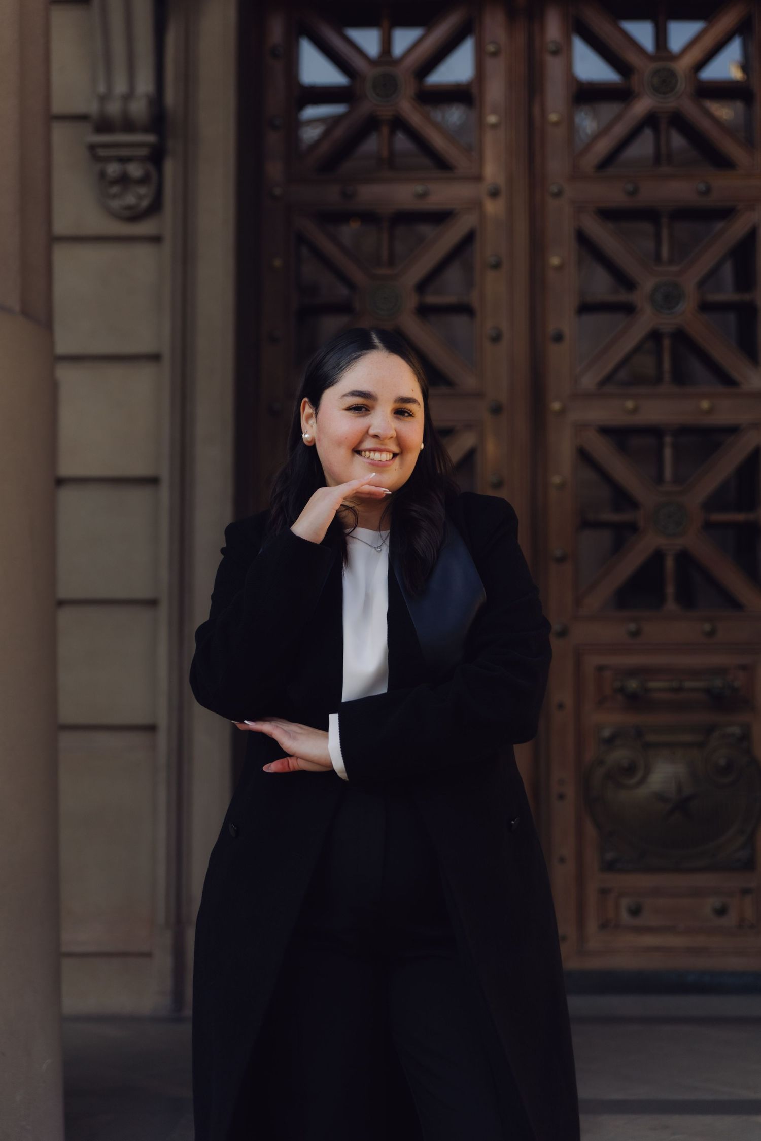 Smiling young woman in black coat leaning against historic building with decorative wooden doors.