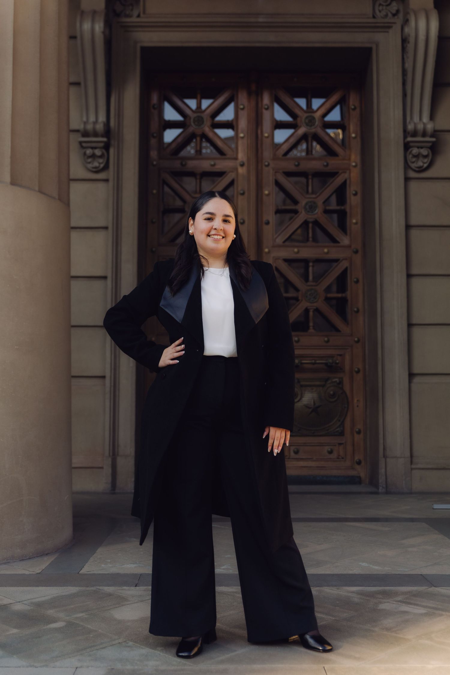 Young woman in black graduation gown and wide-leg pants standing confidently outside ornate building.
