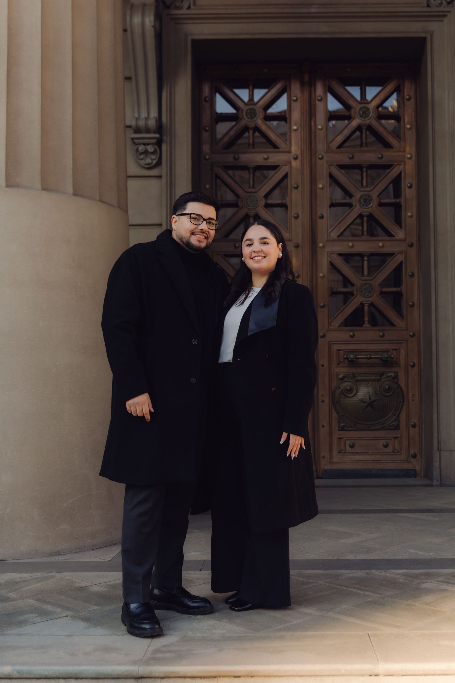 Smiling couple dressed in formal black attire posing together outside ornate wooden building doors.