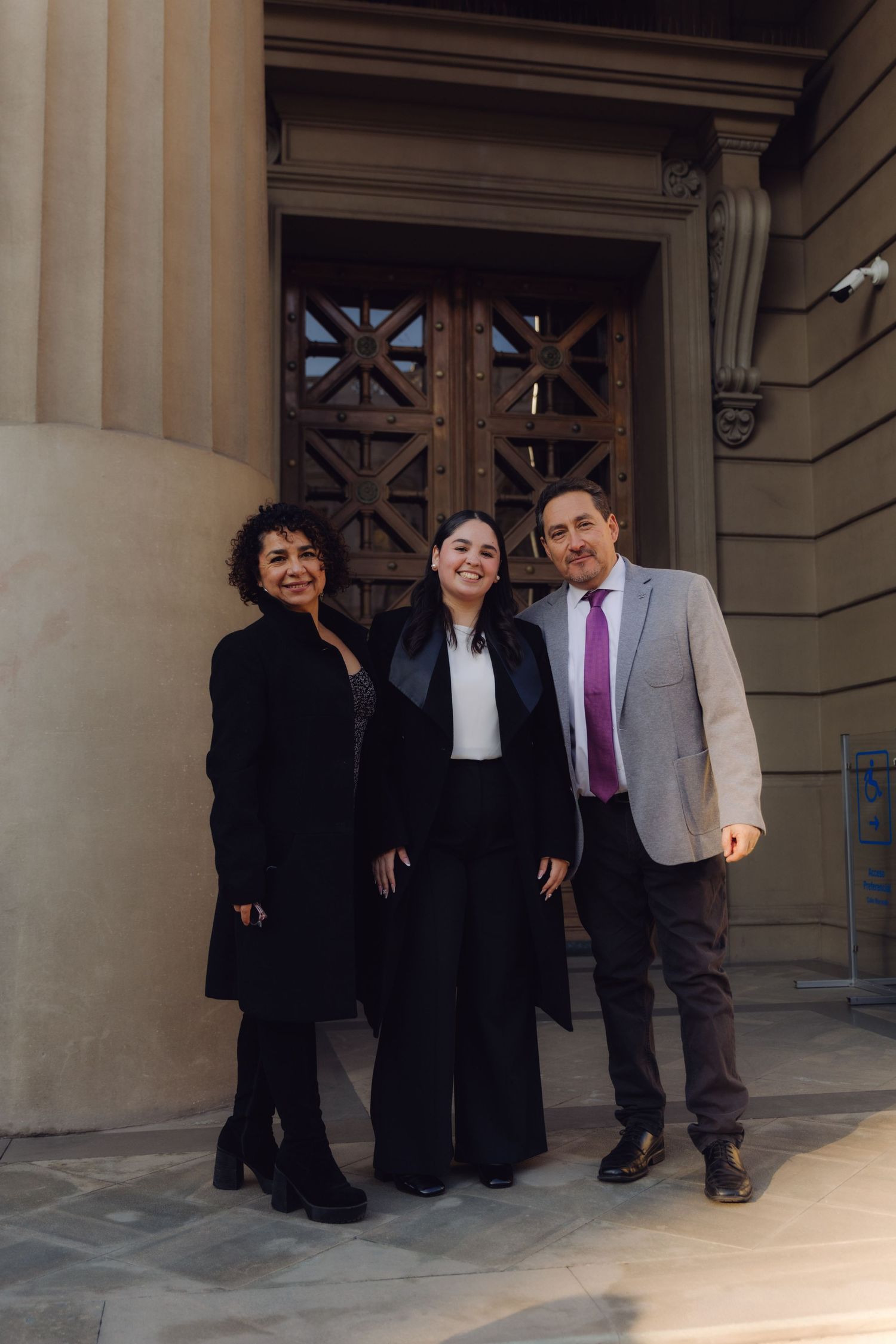 Graduate in black gown celebrating with proud parents outside historic building entrance.