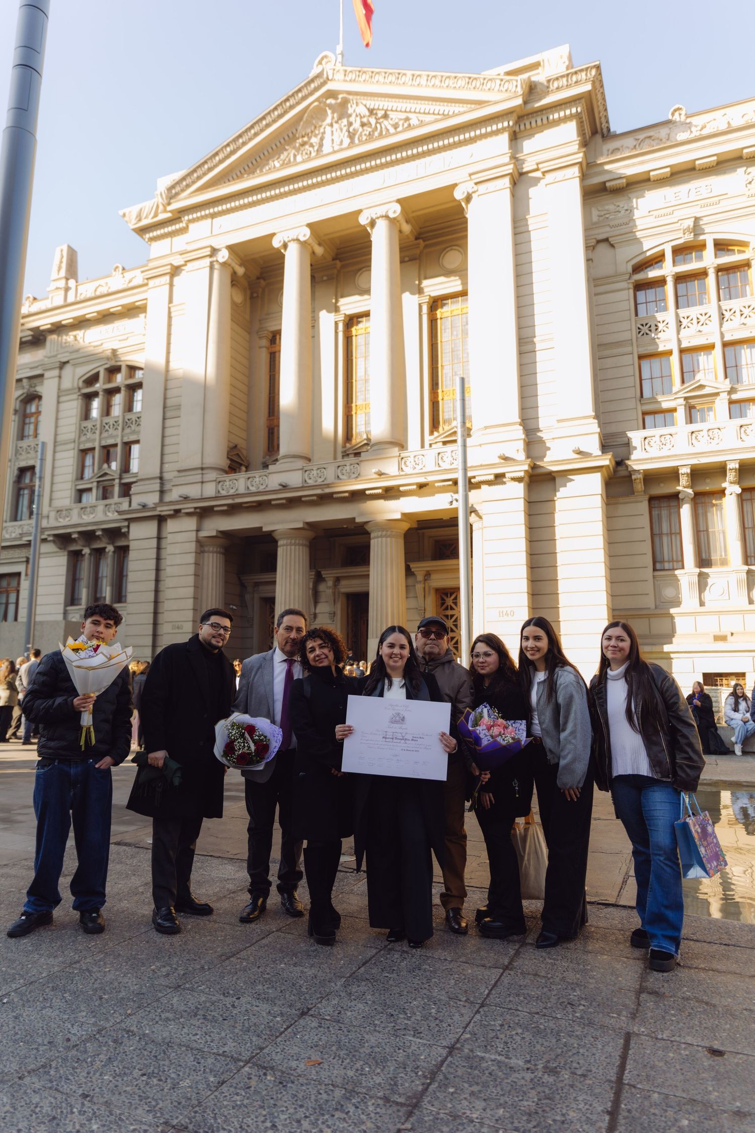 Group of graduates celebrating outside a neoclassical university building, holding diplomas and flowers.