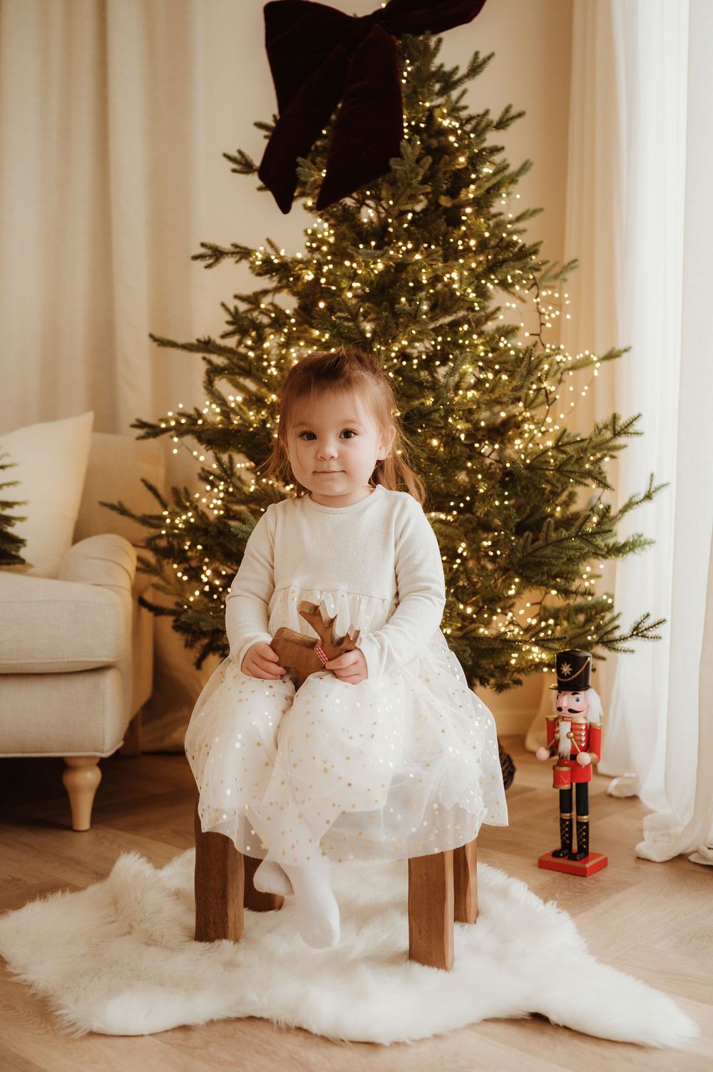 Young girl in white dress sits on stool before lit Christmas tree with nutcracker, holding reindeer toy.