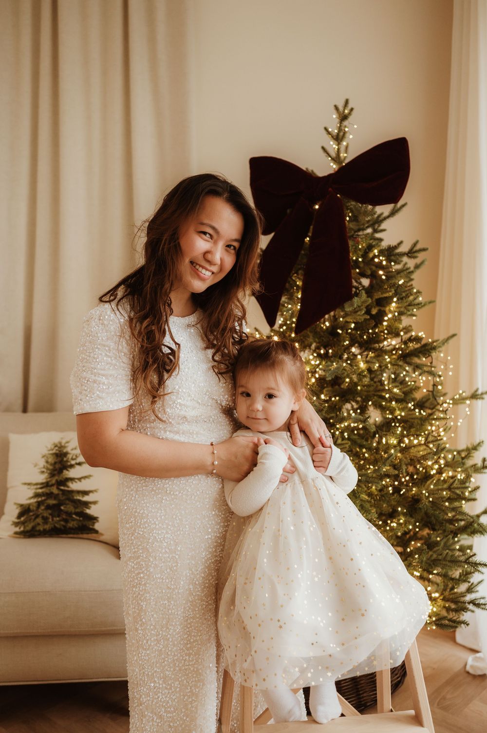 Mother and baby girl in matching white dresses pose by a decorated Christmas tree with warm lights.