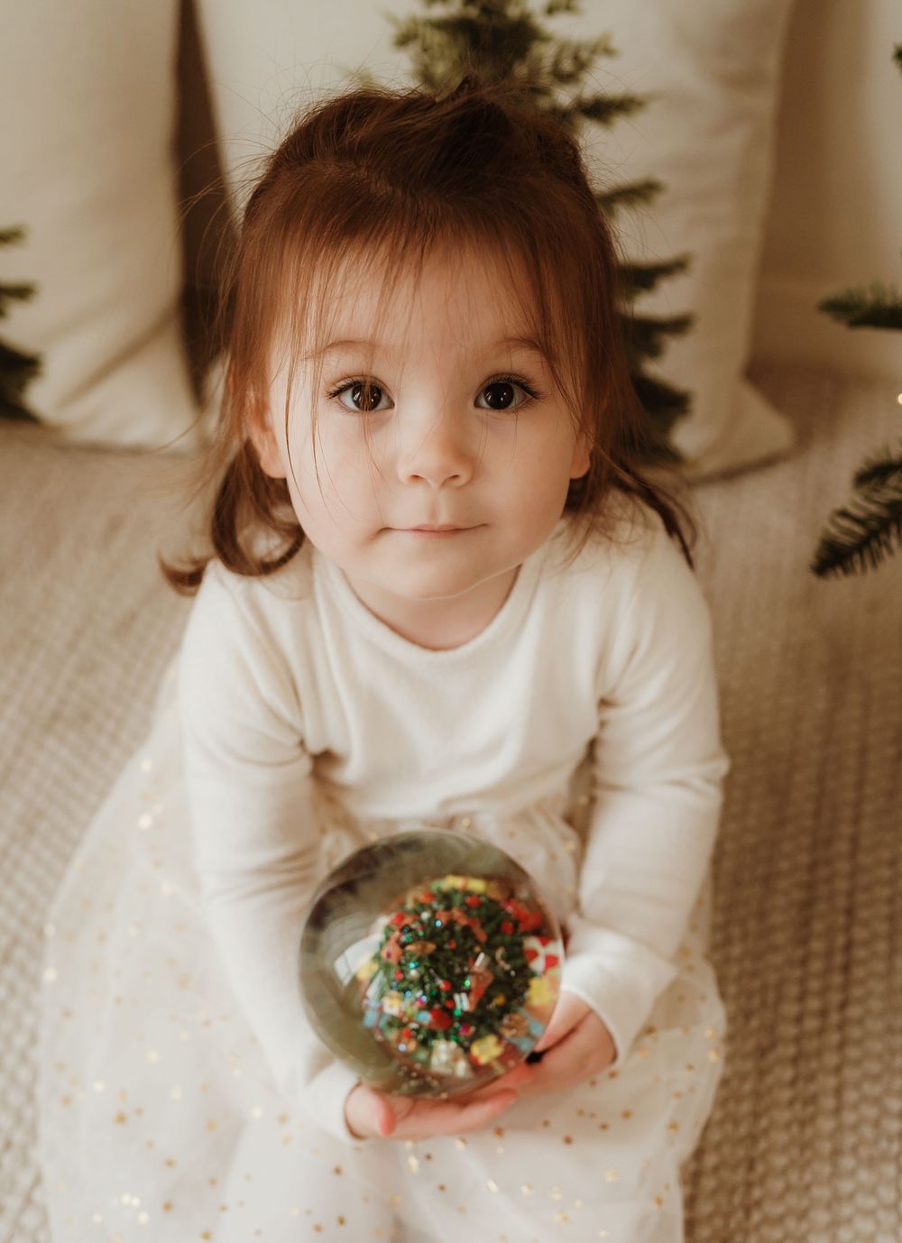 Toddler girl in white dress holding a Christmas snow globe ornament with holiday decorations in background.