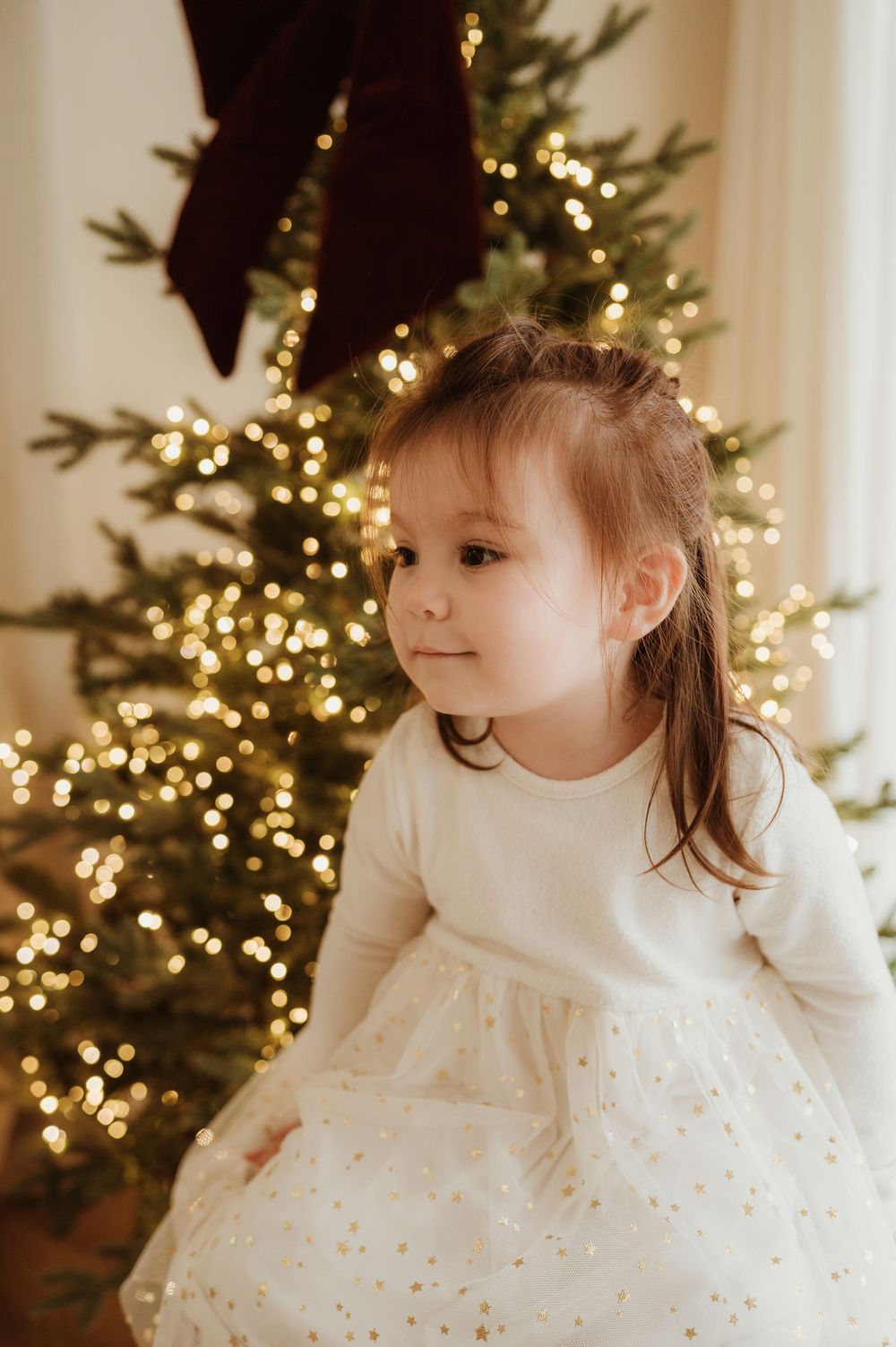 A young girl in a white star-print dress stands before a glowing Christmas tree adorned with warm golden lights.