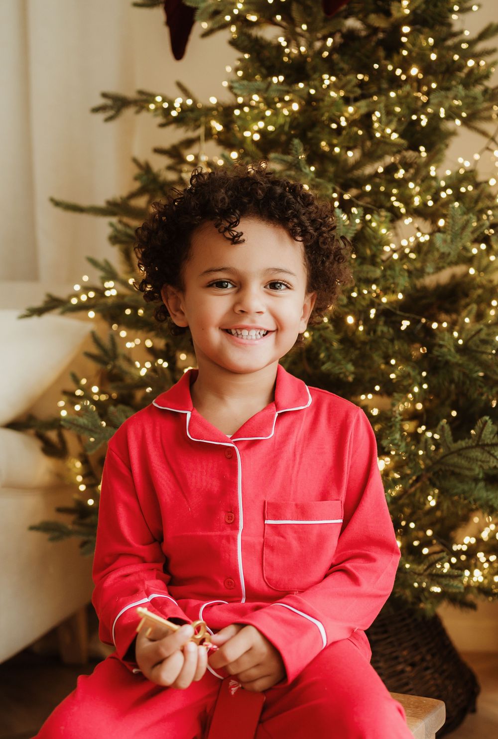 Smiling child in red Christmas pajamas sitting in front of a decorated tree with warm glowing lights.
