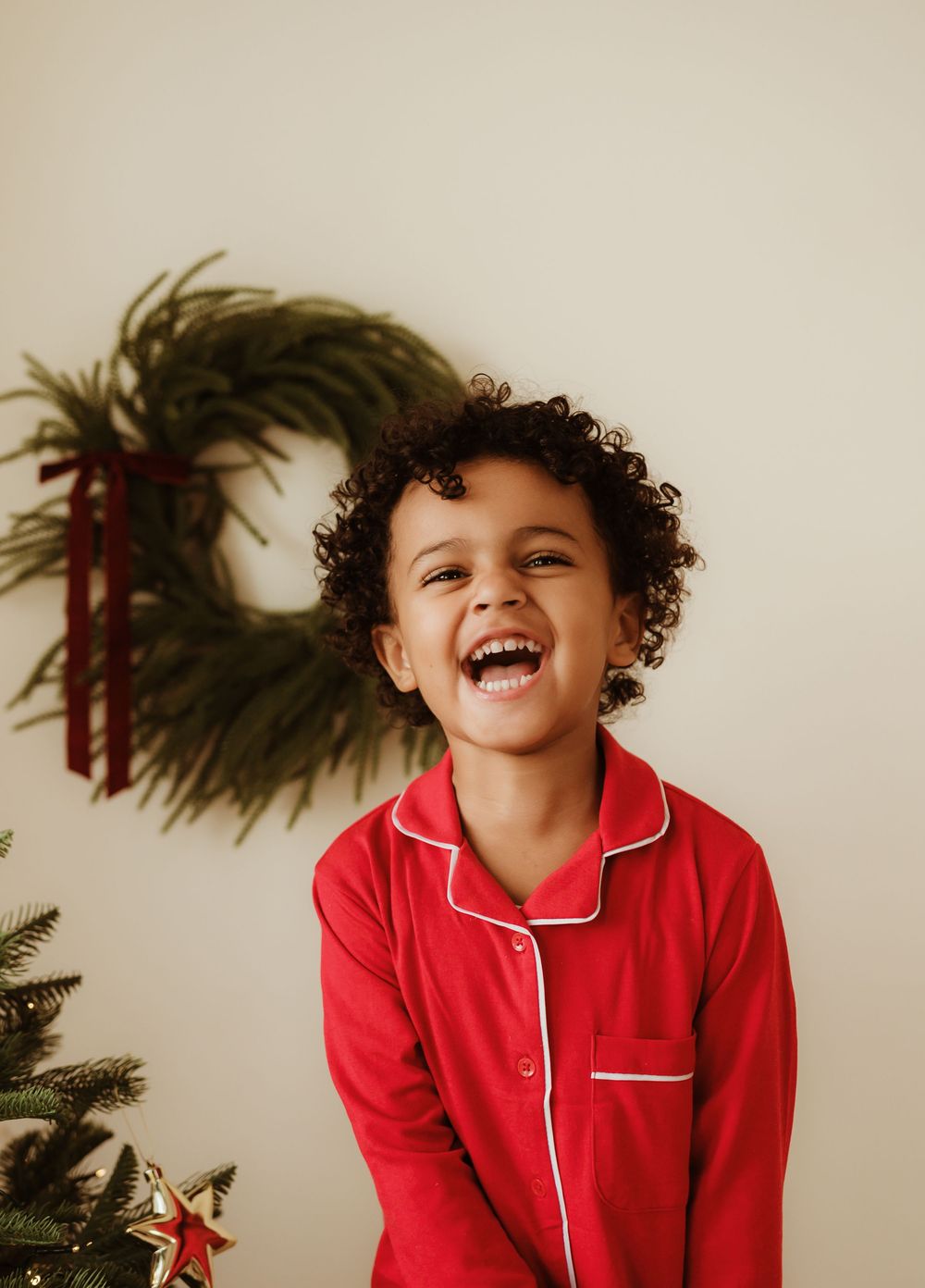 A joyful child in red Christmas pajamas laughs beside a festive wreath and holiday tree.