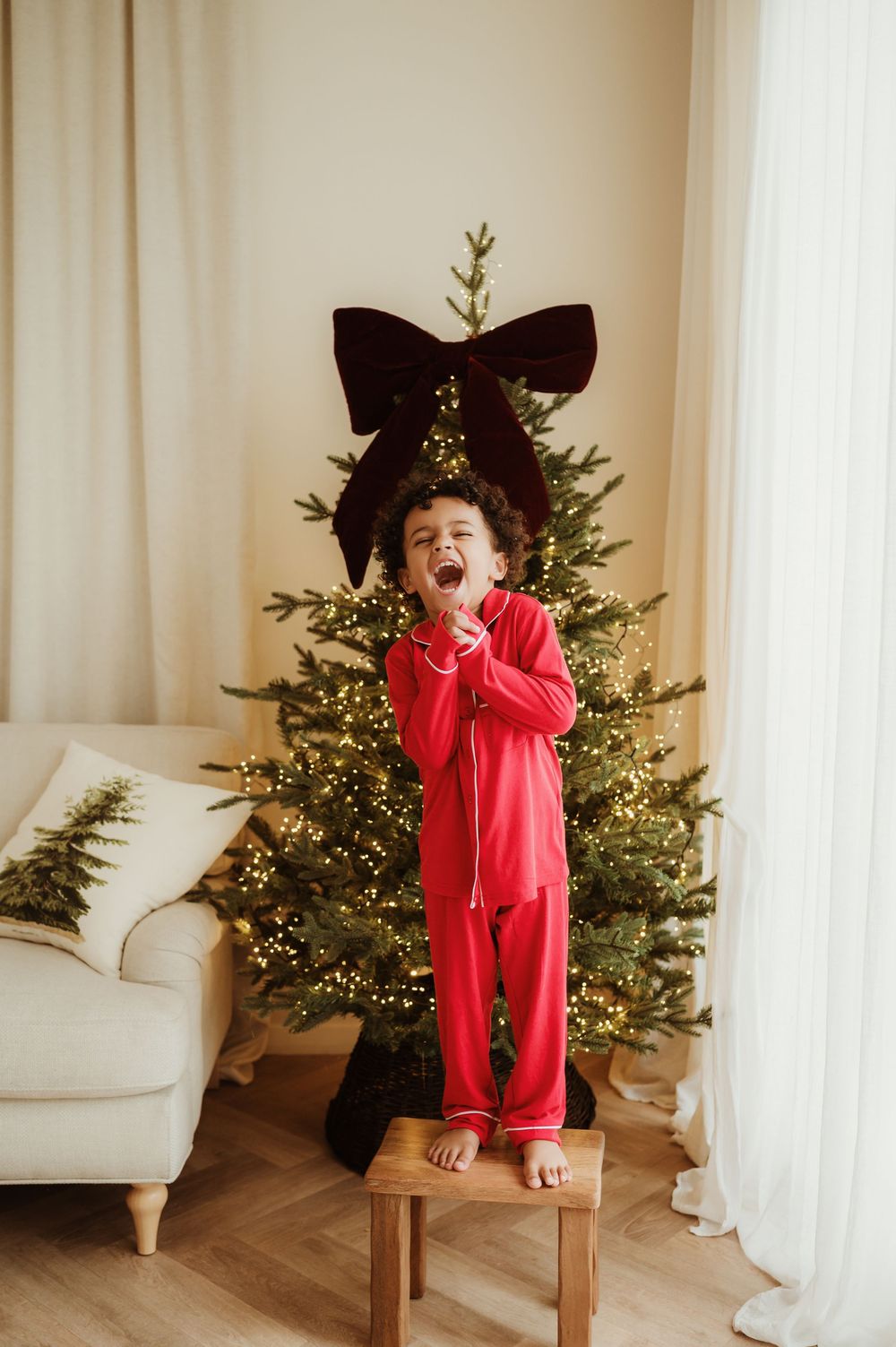 Child in red pajamas standing on a stool in front of a decorated Christmas tree with a velvet bow.