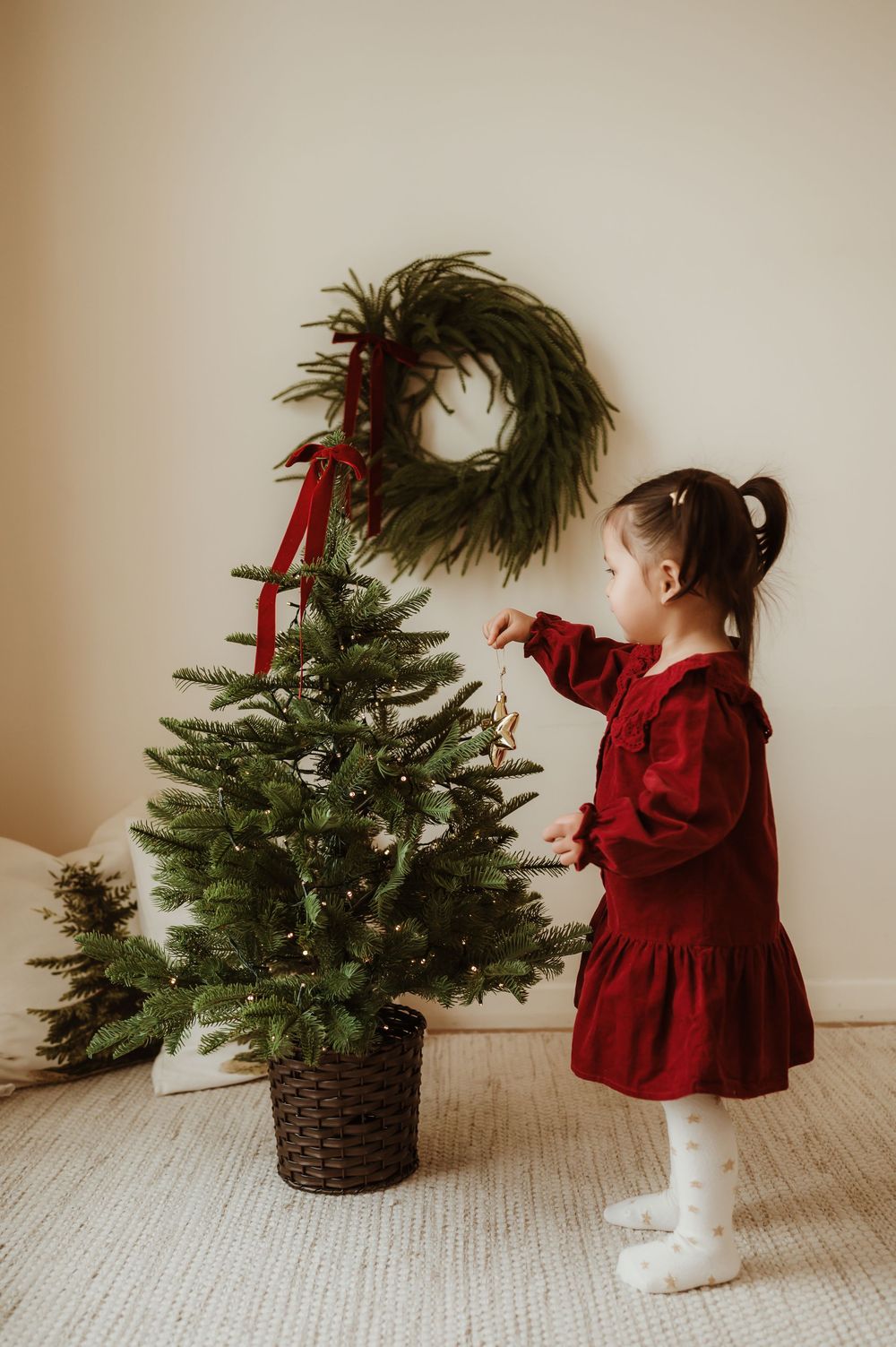 Young girl in red dress decorating a small Christmas tree with ornaments, wreath with red ribbon on wall behind.