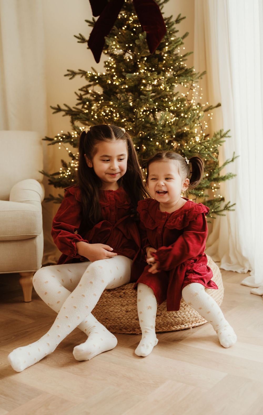 Two young girls in matching red dresses and white tights smile joyfully in front of a decorated Christmas tree.