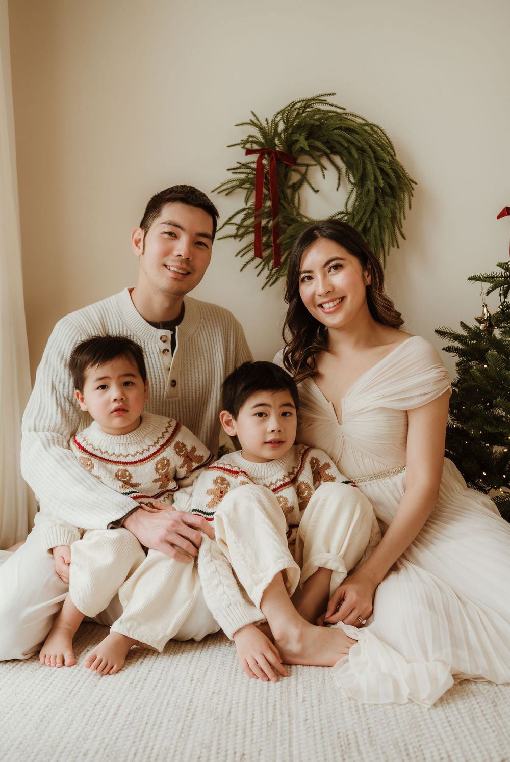 Family of four in matching cream and gingerbread outfits posing for a Christmas portrait with wreath and tree.