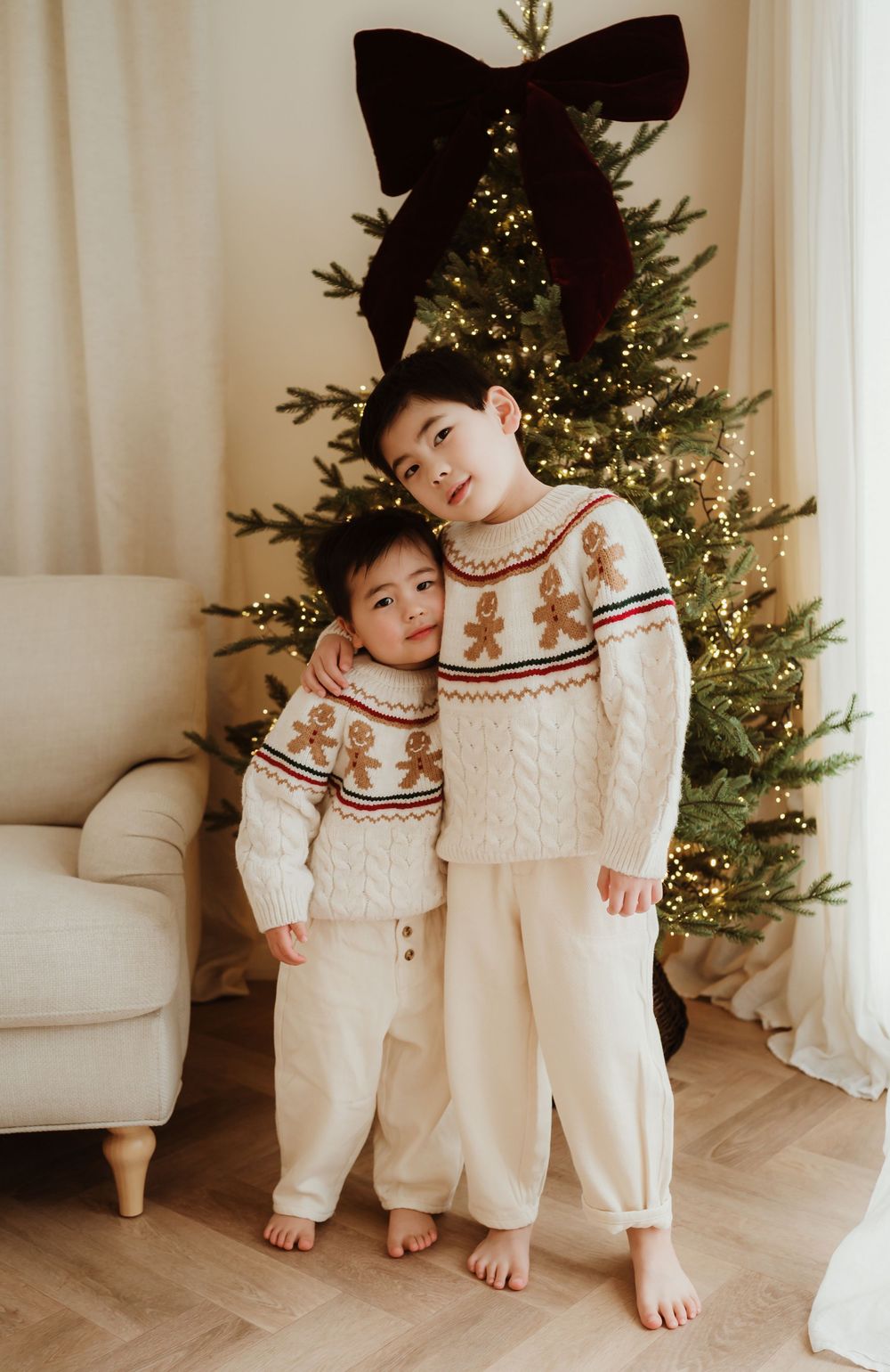 Two children in matching gingerbread sweaters and cream pants pose in front of a decorated Christmas tree.