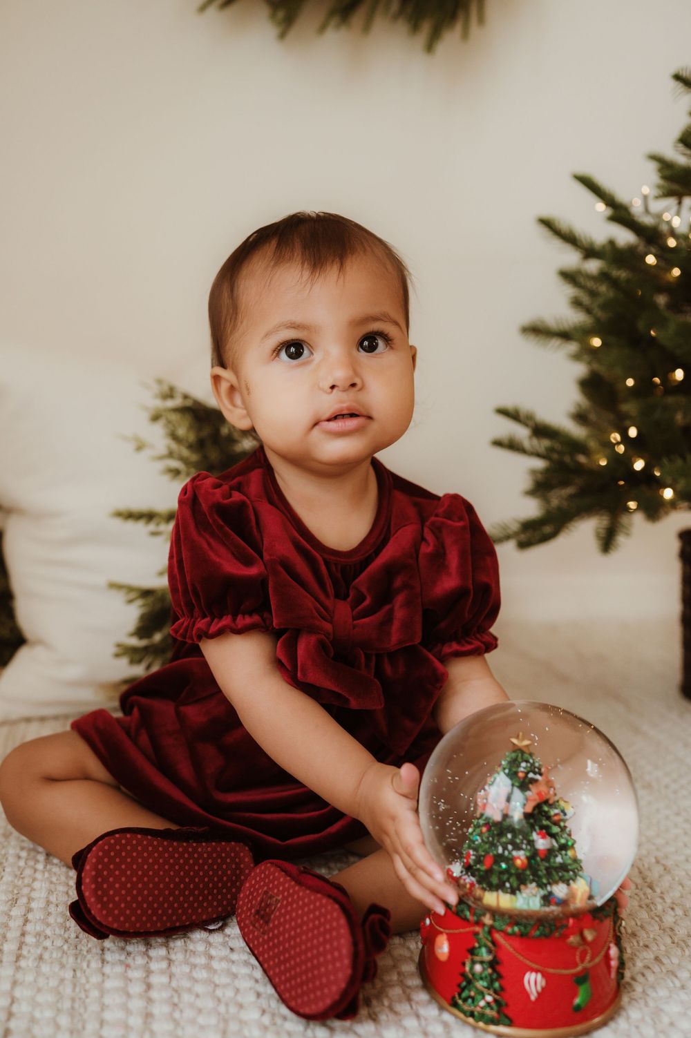 Baby girl in red velvet dress holds Christmas snow globe beside decorated tree.