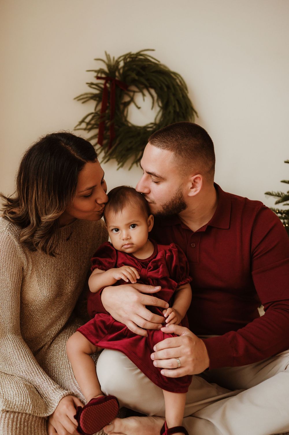 A young family shares a tender Christmas moment, parents kissing their baby girl in a red dress near a holiday wreath.