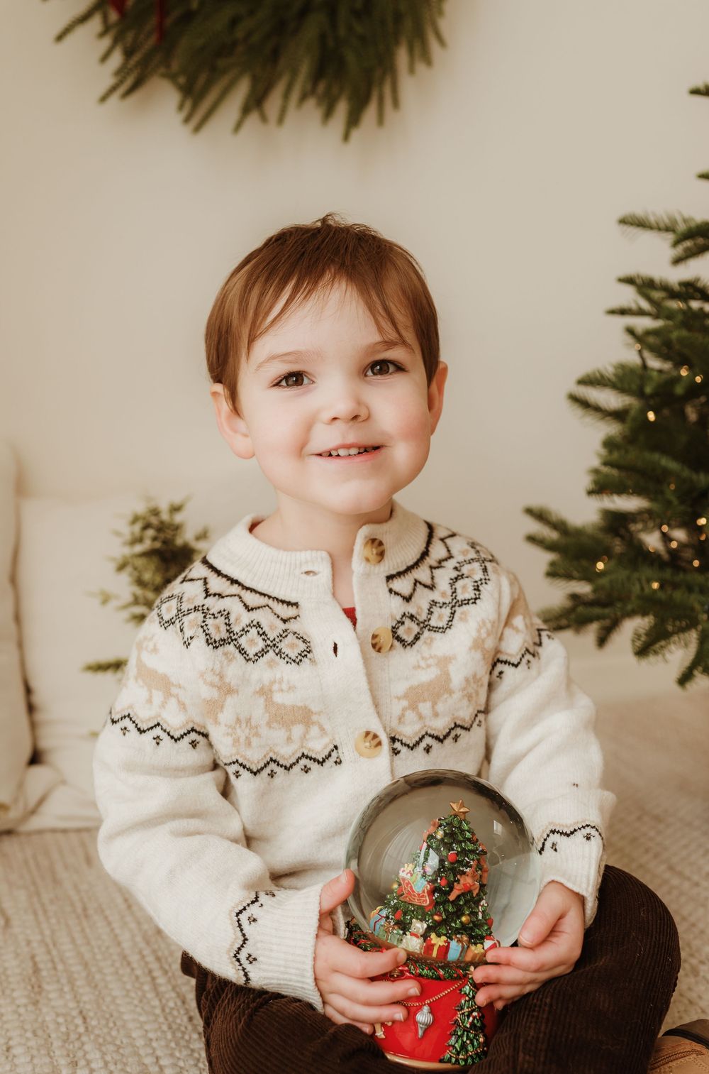 Young child in reindeer sweater holding a Christmas snow globe, sitting near a decorated holiday tree.