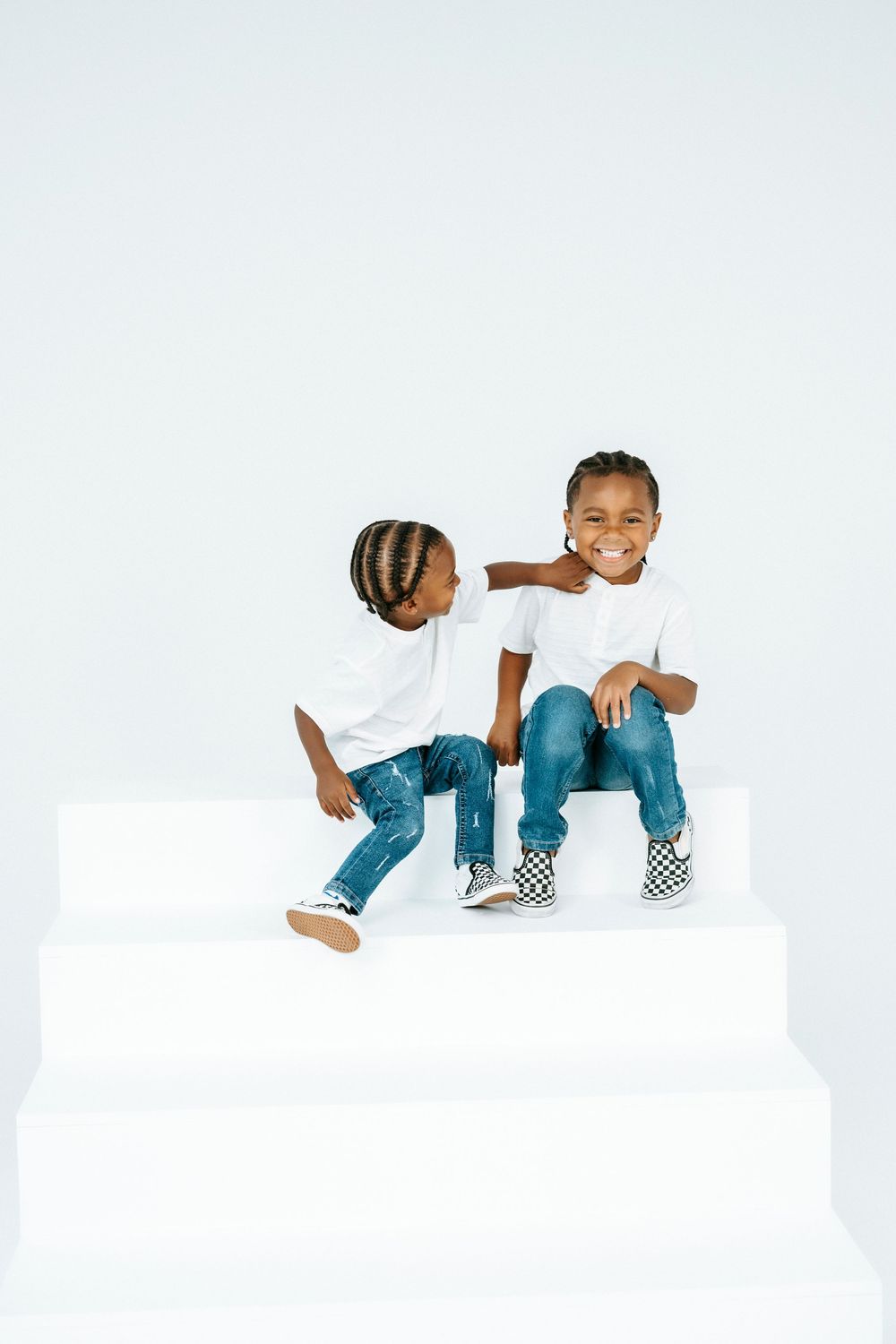 Two young African American boys in white tees and jeans sit on white steps, smiling and posing together.
