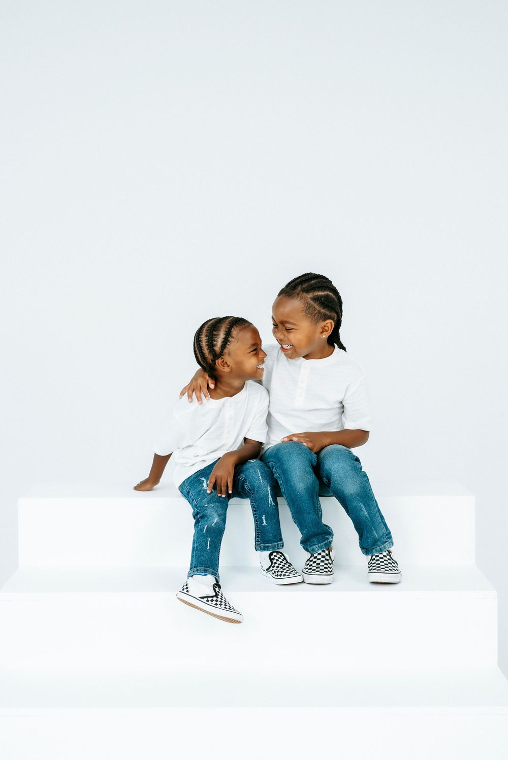 Two young African American children in white shirts and jeans sitting together smiling on a white block.