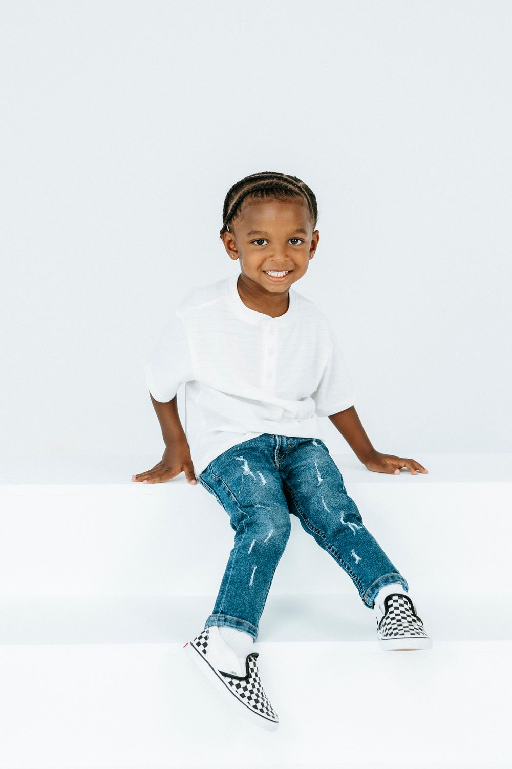 Smiling young boy in white shirt, ripped jeans, and checkered Vans sneakers sitting on white surface.