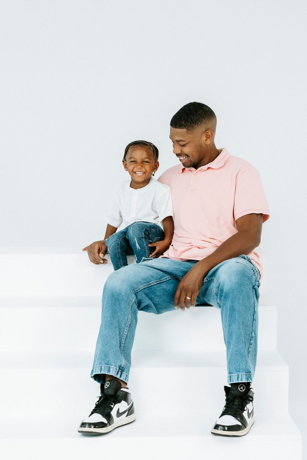 A smiling father and young son sit together on white steps, both wearing jeans and sneakers in a studio setting.