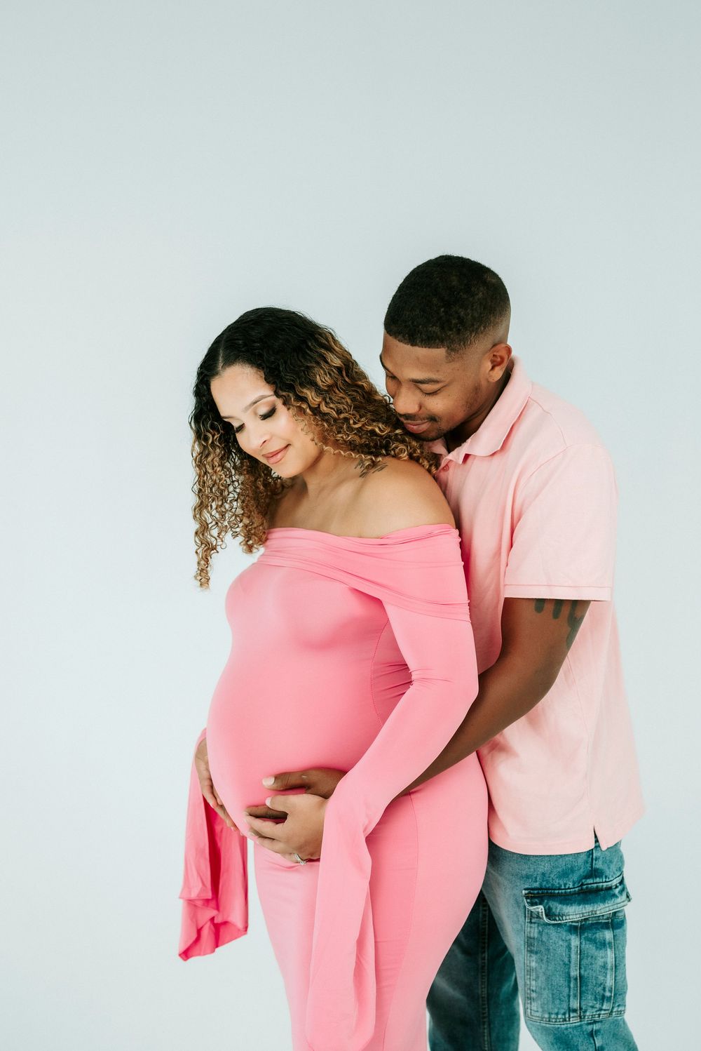 Couple in maternity photoshoot, woman in pink off-shoulder dress cradling baby bump, man embracing her from behind.