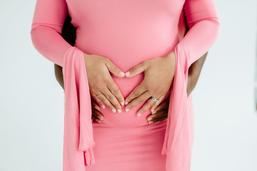 Pregnant woman in pink dress forming heart shape with hands on baby bump, maternity photo.